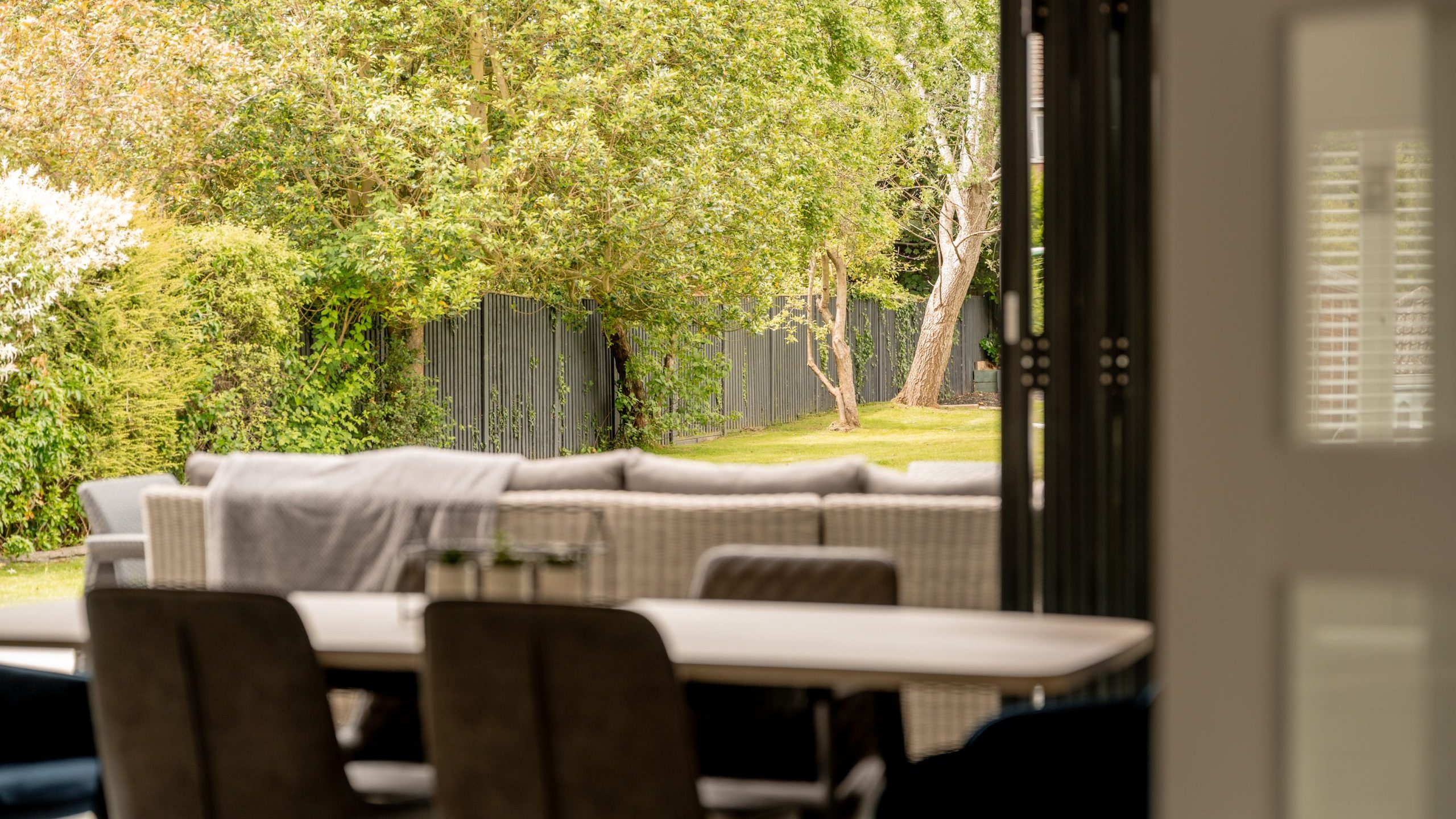 A modern dining area opens onto a patio with outdoor seating, overlooking a lush green garden bordered by trees and a dark wooden fence. The space is bright and inviting.