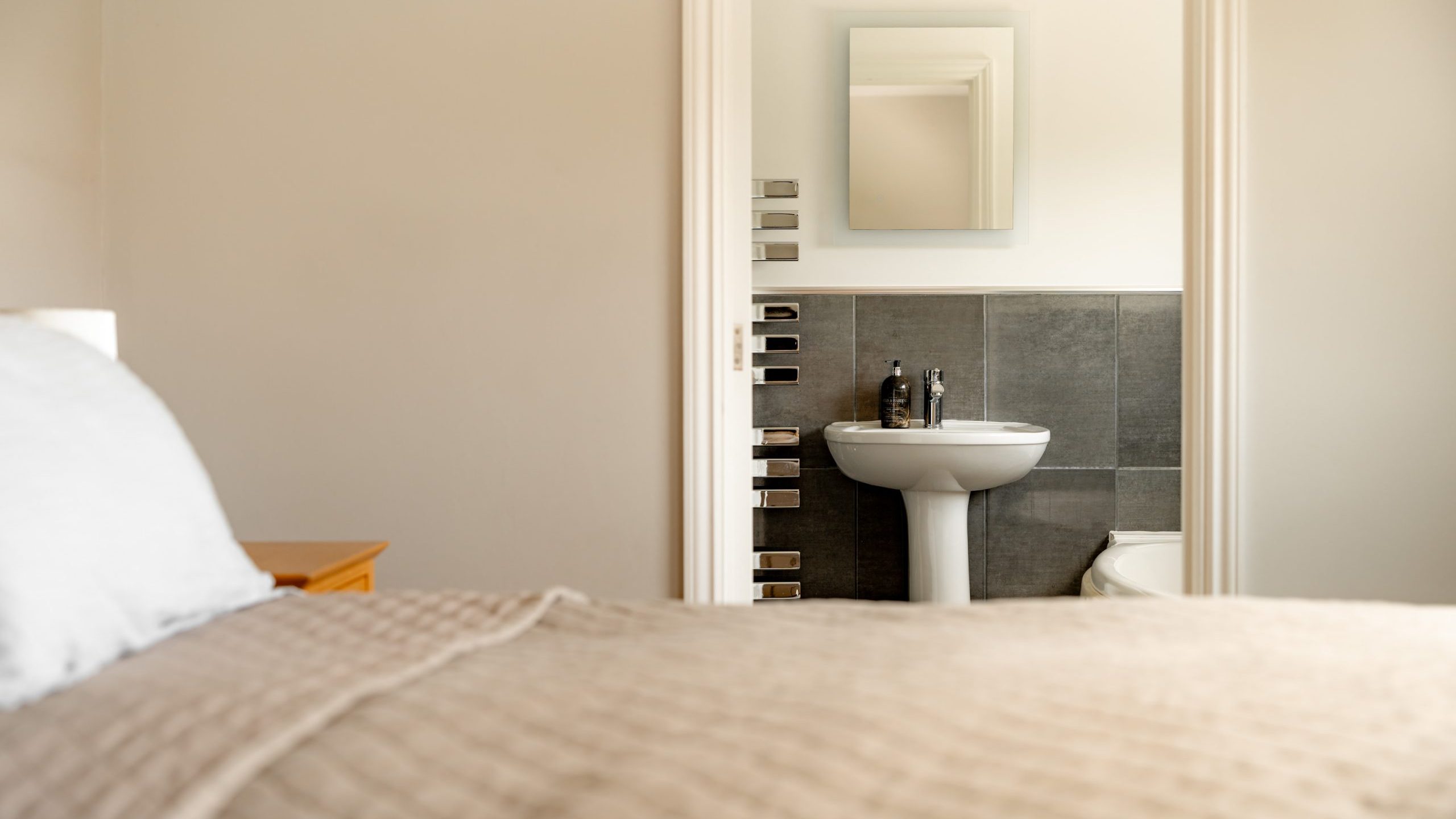 A tidy bedroom with a beige quilt on the bed and a view into an adjoining bathroom, featuring a white pedestal sink, a mirror above it, and gray tiled walls.