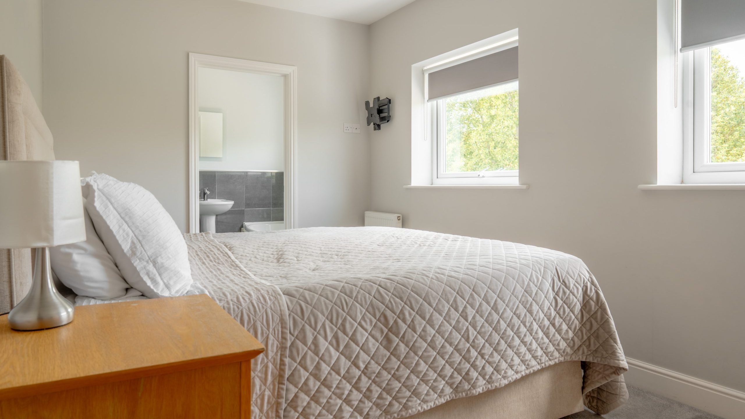 A bright, tidy bedroom with a double bed, beige quilt, wooden nightstand, lamp, and two windows with gray blinds. An open door reveals a bathroom with a sink and mirror. The room has light-colored walls and carpet.