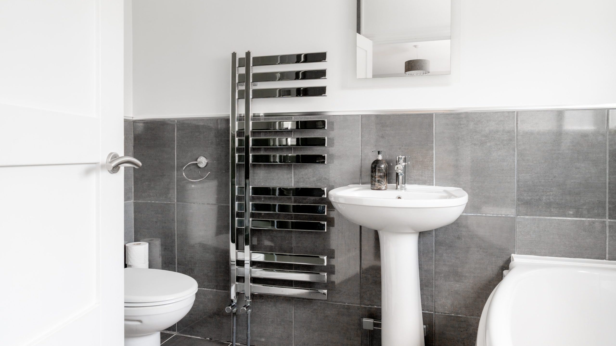 Modern bathroom with gray tiled walls, a white toilet, pedestal sink, rectangular mirror, stainless steel towel radiator, and a bathtub partially visible on the right. Soap dispenser and bottle are on the sink.