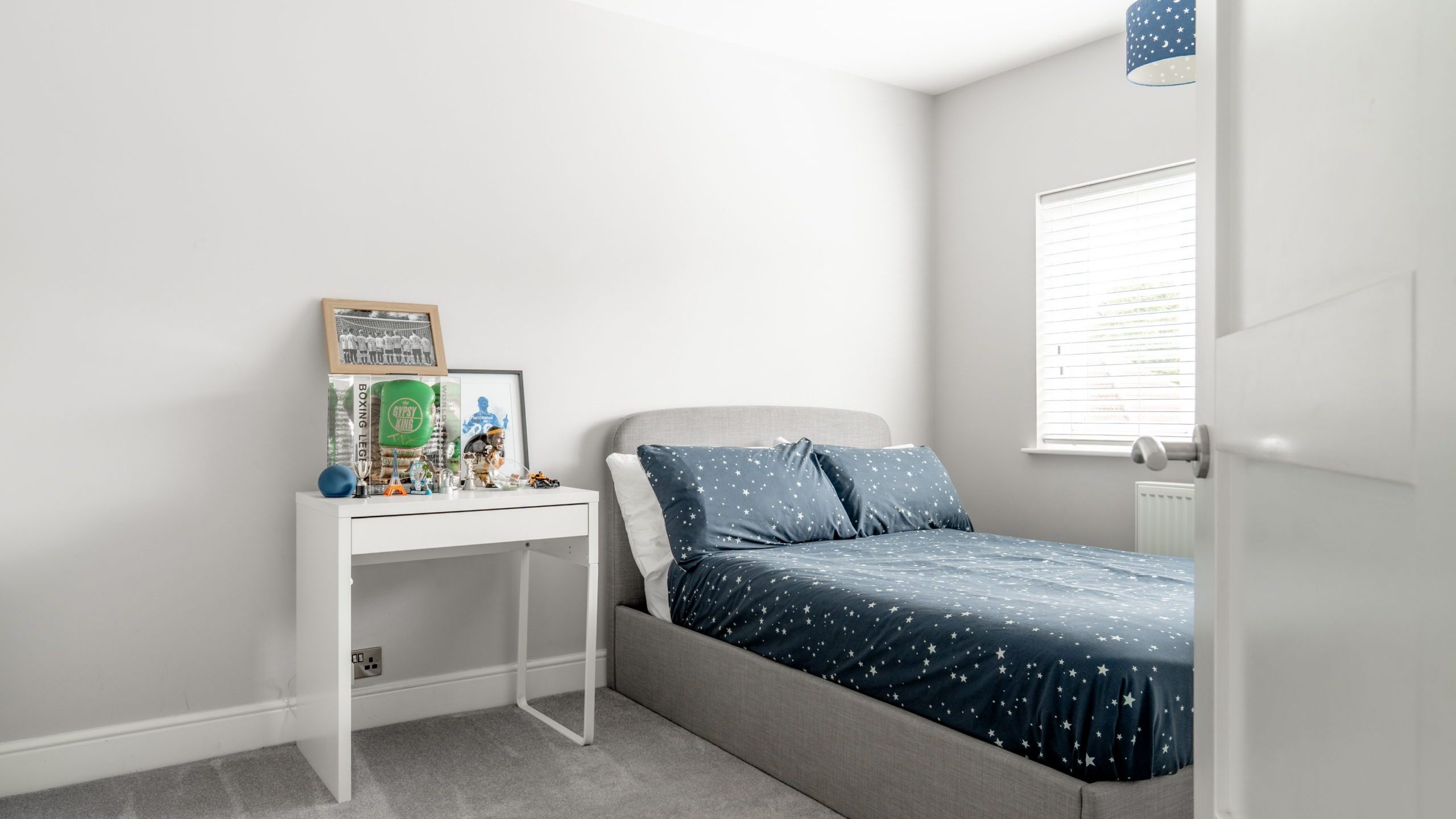 A modern, minimal bedroom with a grey bed, blue star-patterned bedding, a small white desk with toys and framed pictures, grey carpet, and a window with white blinds letting in natural light.
