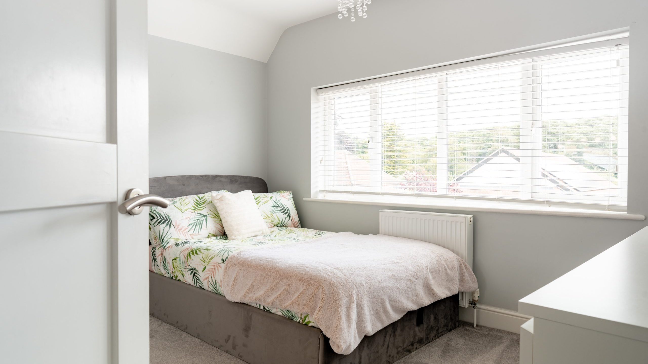 A bright, modern bedroom with a large window, double bed with tropical-patterned bedding, light gray walls, beige carpet, and a white chest of drawers. Natural light fills the space.