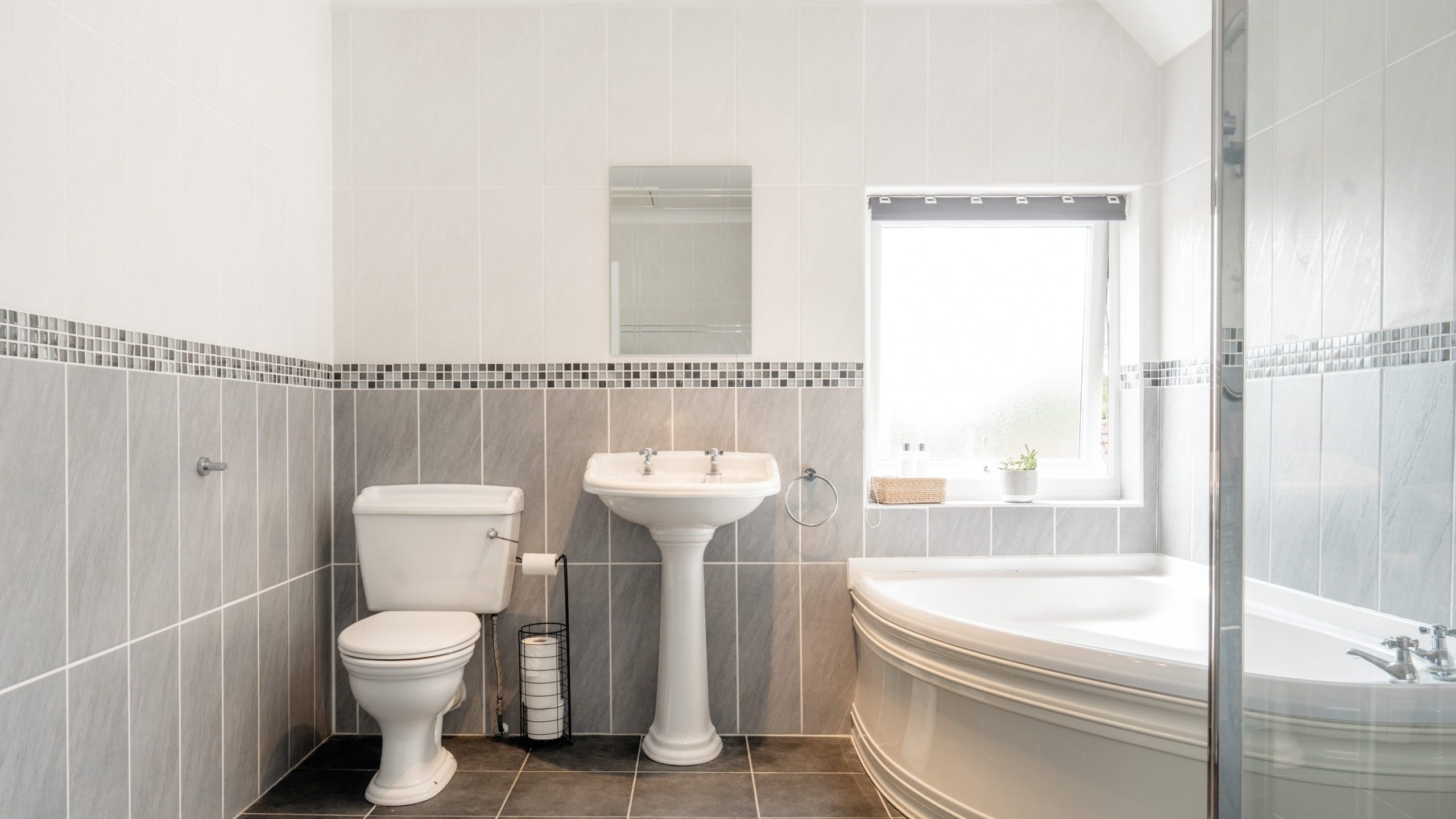 Modern bathroom with gray and white tiles, featuring a toilet, pedestal sink with a mirror above, large corner bathtub, window with blinds, and a small plant on the windowsill.