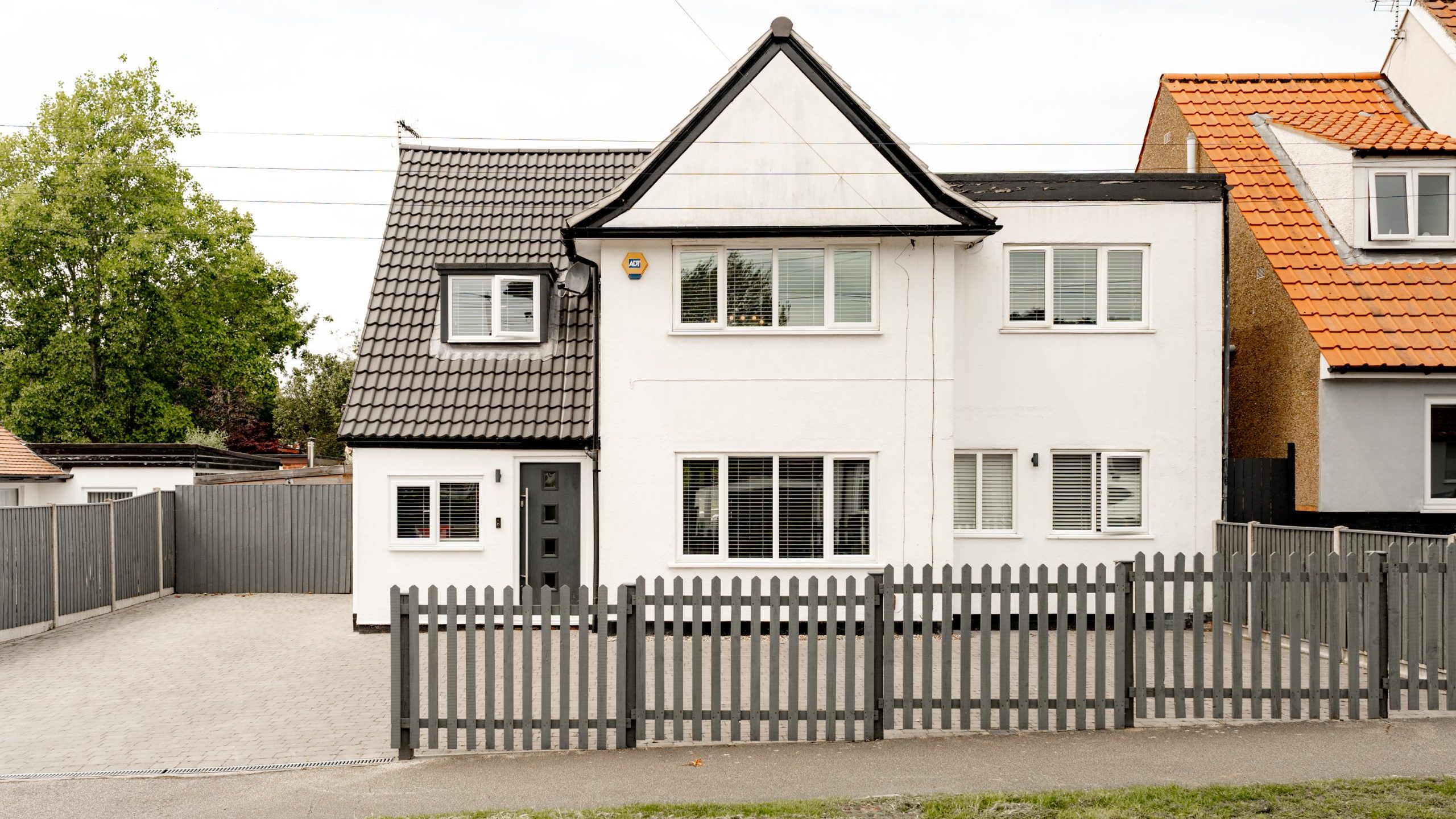 A modern two-story white house with black roof tiles, several windows, and a gray front door, surrounded by a gray picket fence and driveway, set on a residential street.