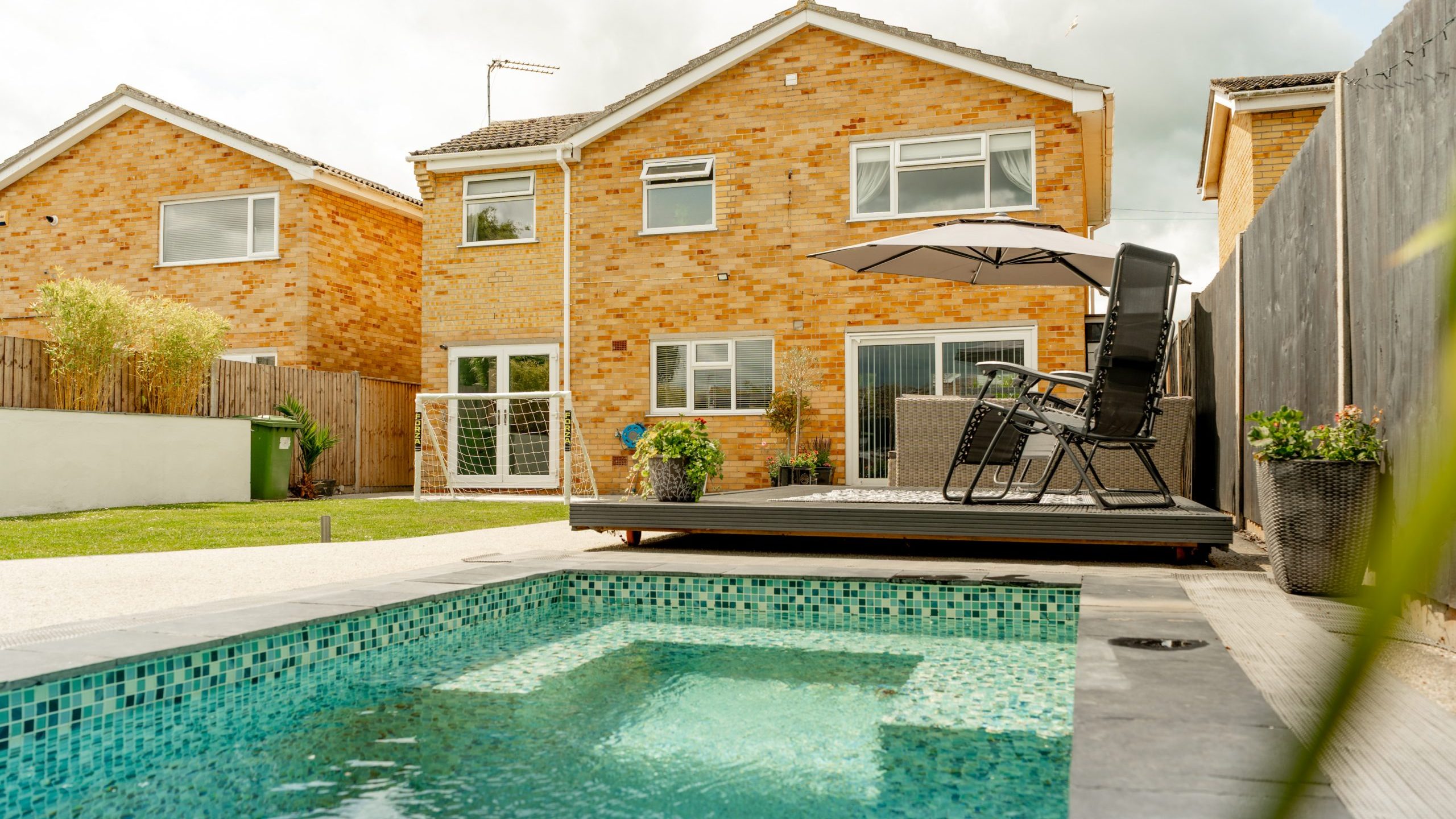 Backyard view of a modern house with a small swimming pool in the foreground, a tiled patio with lounge chairs under a large umbrella, and neatly manicured grass.