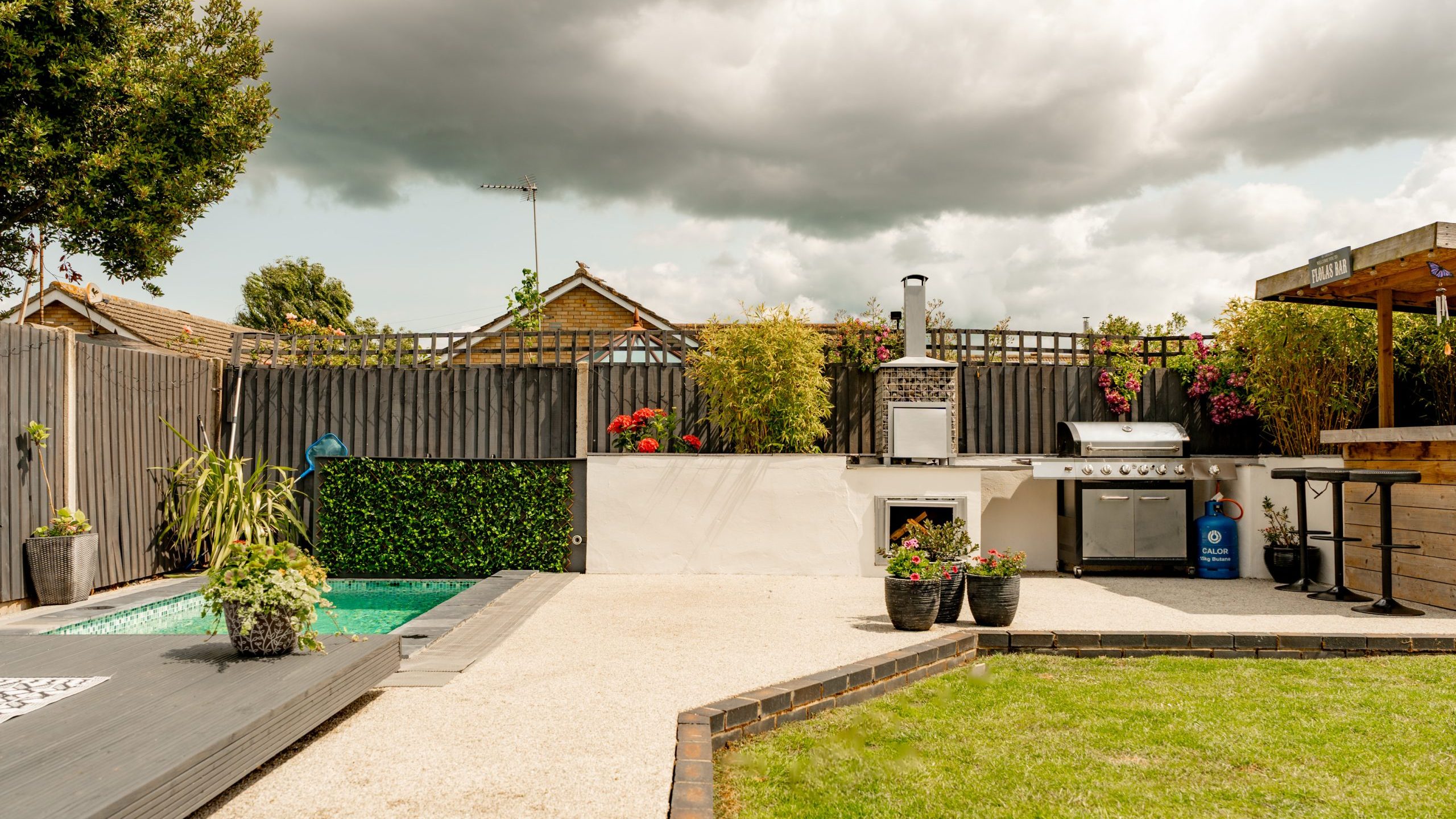A modern backyard with a small pool, outdoor kitchen with a pizza oven and grill, potted plants, a wooden bar area, and a cloudy sky overhead.