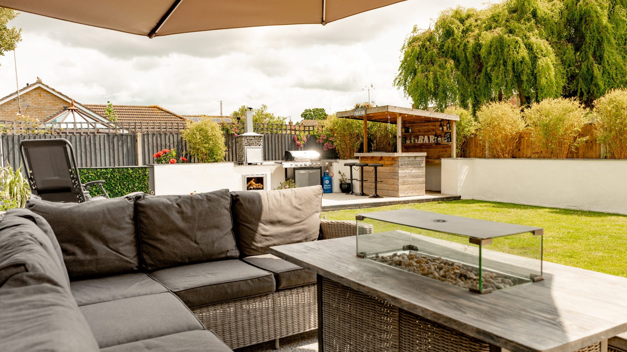 A modern backyard patio with a gray outdoor sofa, a glass-topped fire pit table, a large umbrella, a green lawn, and a wooden outdoor bar surrounded by trees and plants.