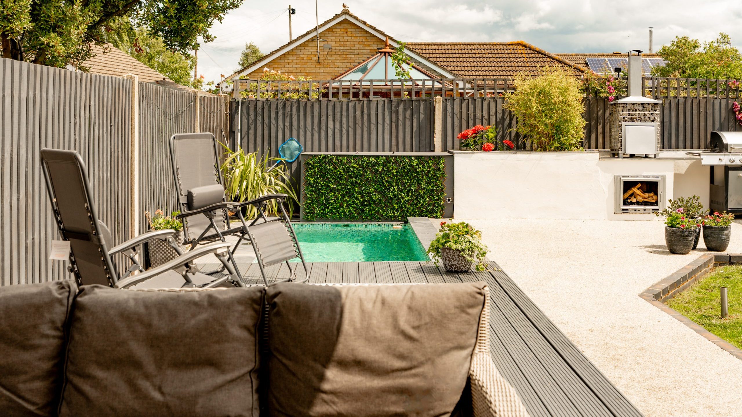 A modern backyard with a small rectangular pool, black lounge chairs, grey cushioned sofa, potted plants, and an outdoor kitchen area with a fireplace and oven, enclosed by a dark wooden fence.