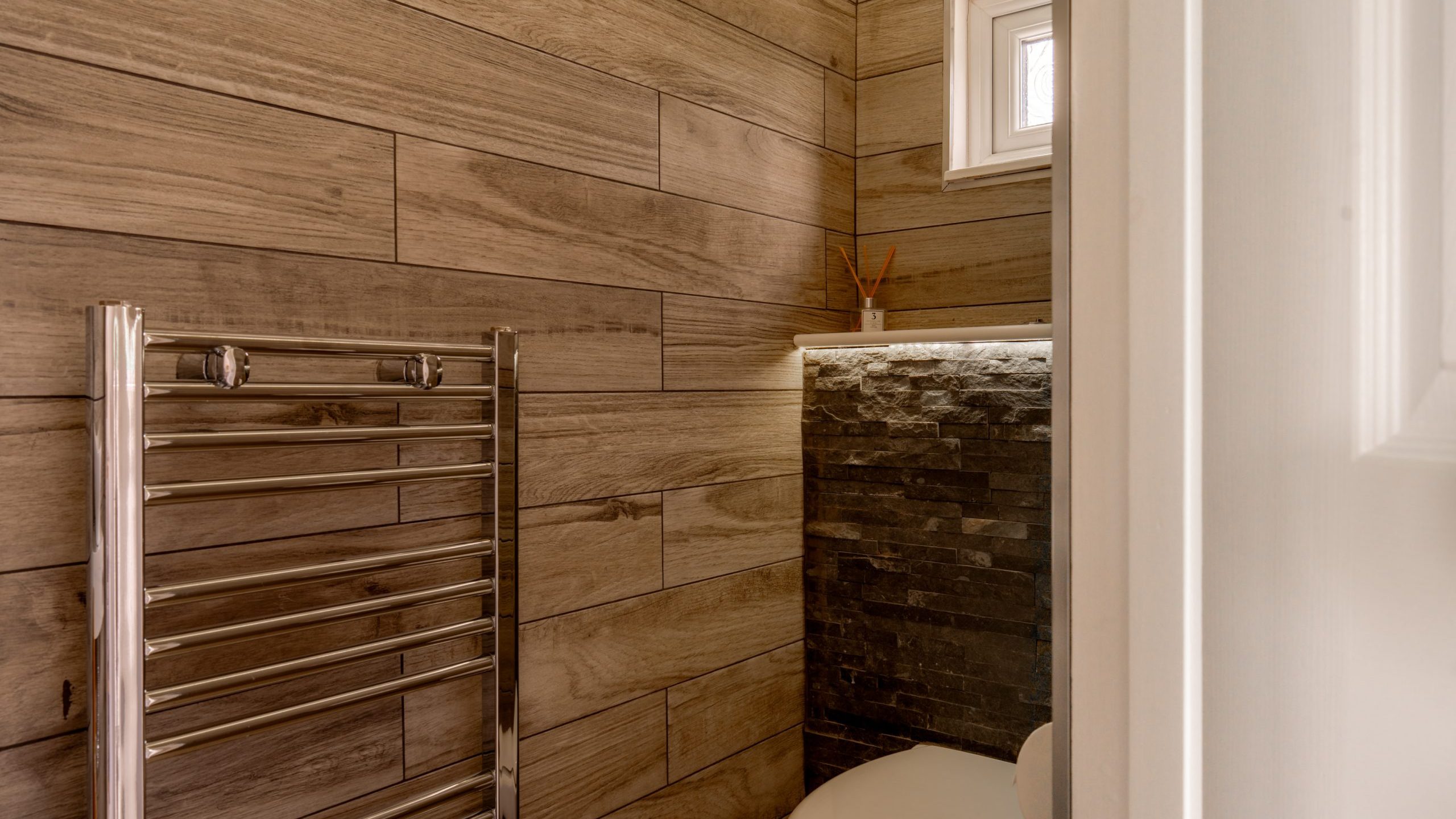 A modern bathroom with wood-look tile walls, a chrome towel warmer, a toilet, a small window, and a stone accent wall with recessed shelf and diffuser.