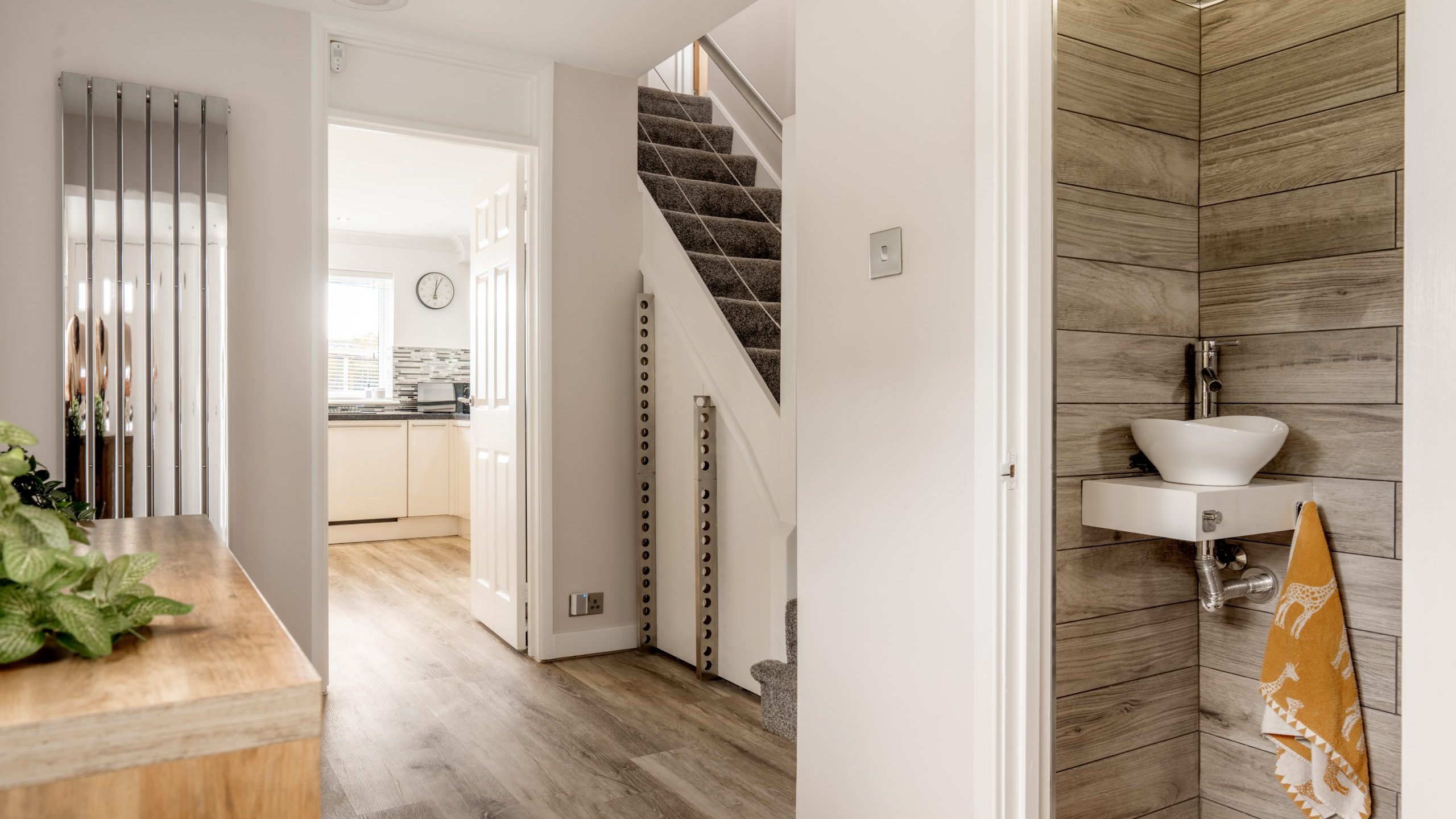Modern hallway with wood flooring, a small sink and towel in a tiled nook, stairs with grey carpet, and a view into a bright kitchen with white cabinets and a wall clock.