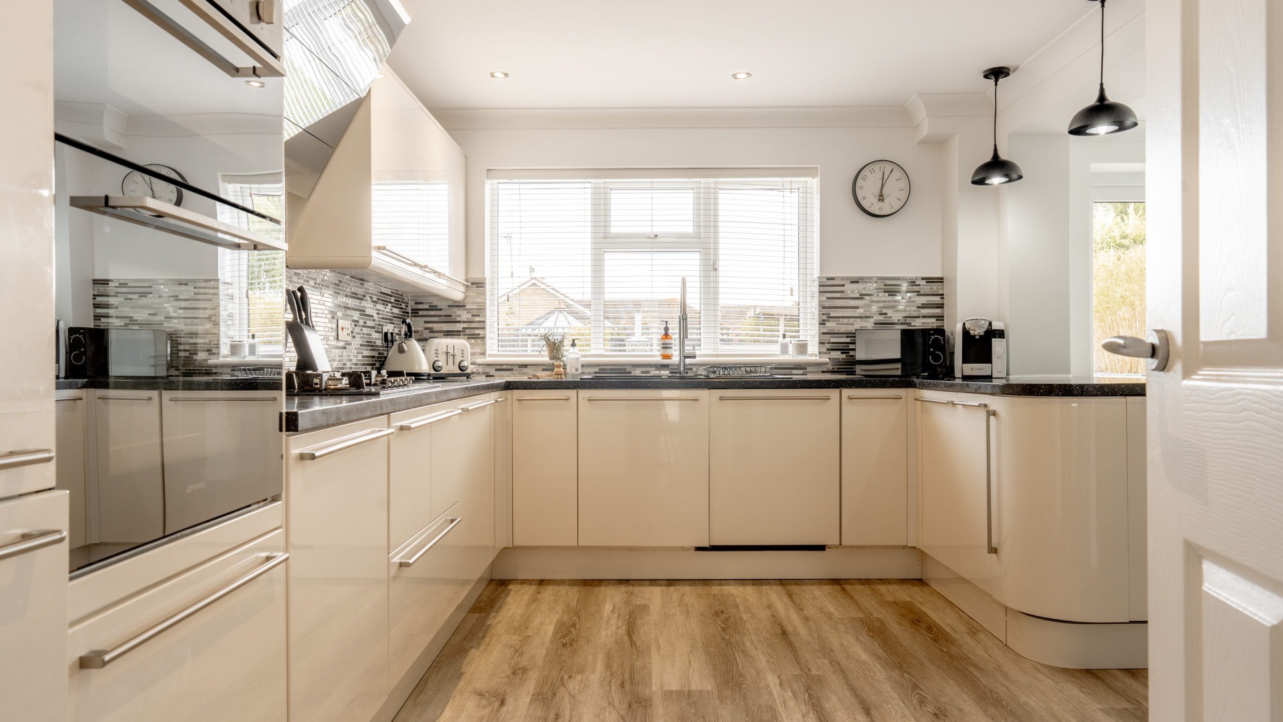 Modern kitchen with glossy white cabinets, built-in appliances, wood-look flooring, black countertops, a large window with blinds, a wall clock, and two hanging pendant lights over the counter.