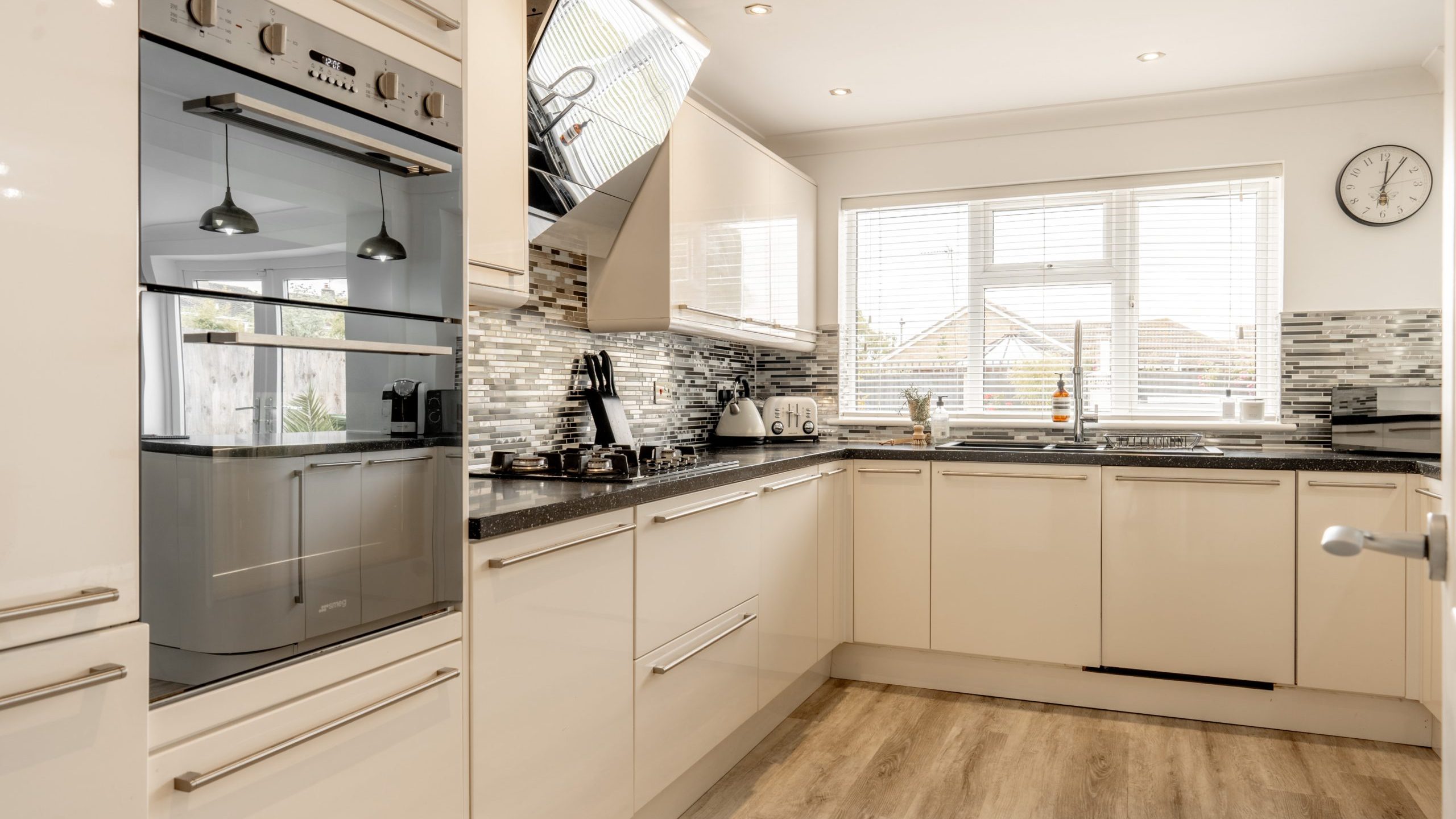 Modern kitchen with glossy cream cabinets, built-in double oven, induction stove, and stainless steel range hood. The backsplash is a mosaic of gray tiles, and there is a large window letting in natural light.