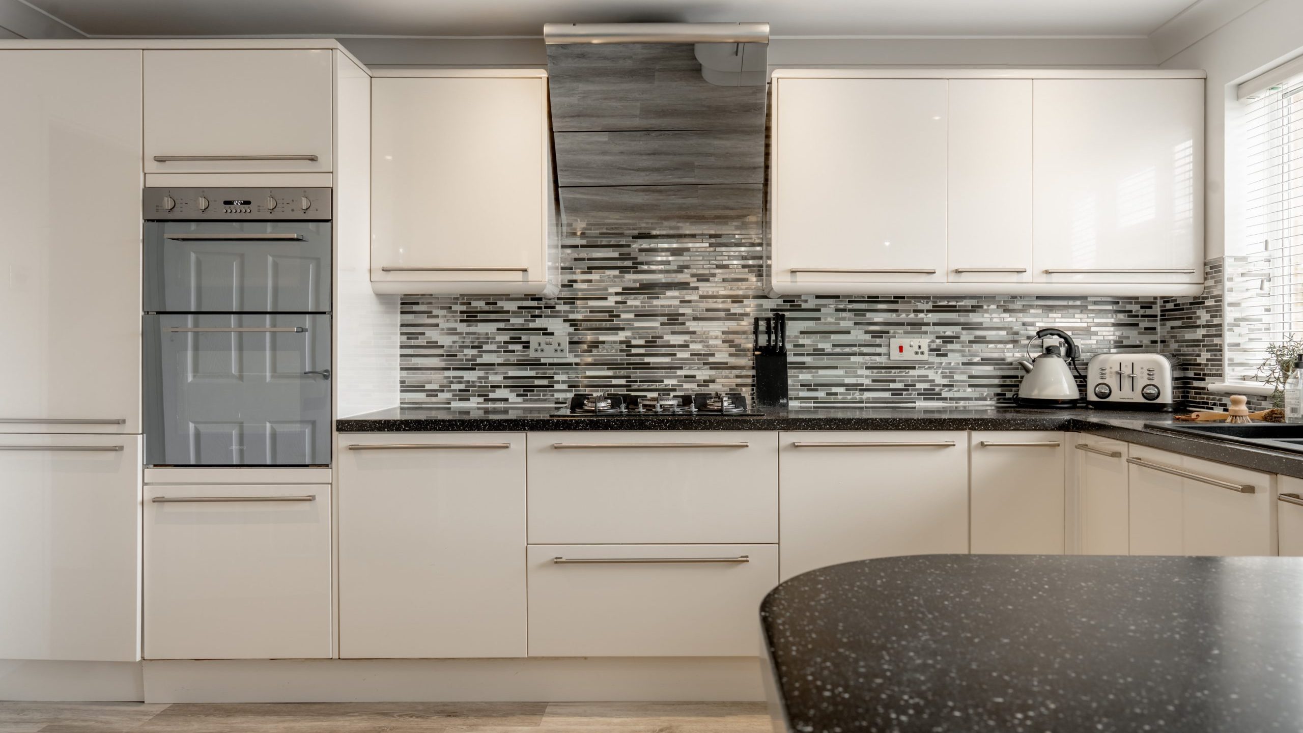 Modern kitchen with white cabinets, a built-in oven, a gas stove, mosaic tile backsplash, black countertop, and small appliances such as a kettle and toaster near a window with blinds.