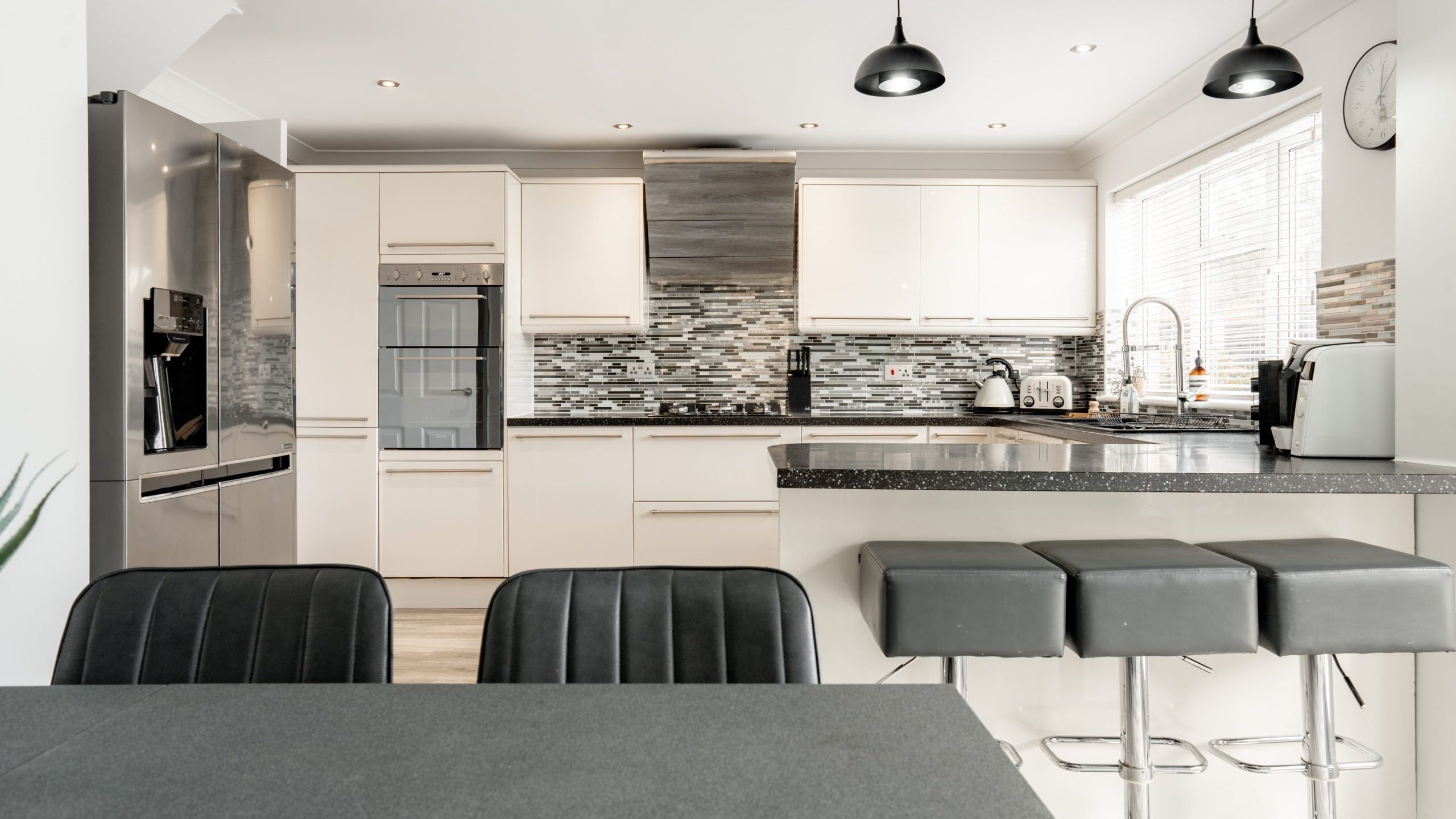 Modern kitchen with white cabinets, stainless steel appliances, black countertop, gray barstools, a dining table with black chairs in the foreground, and a tiled backsplash. Three pendant lights hang above the countertop.