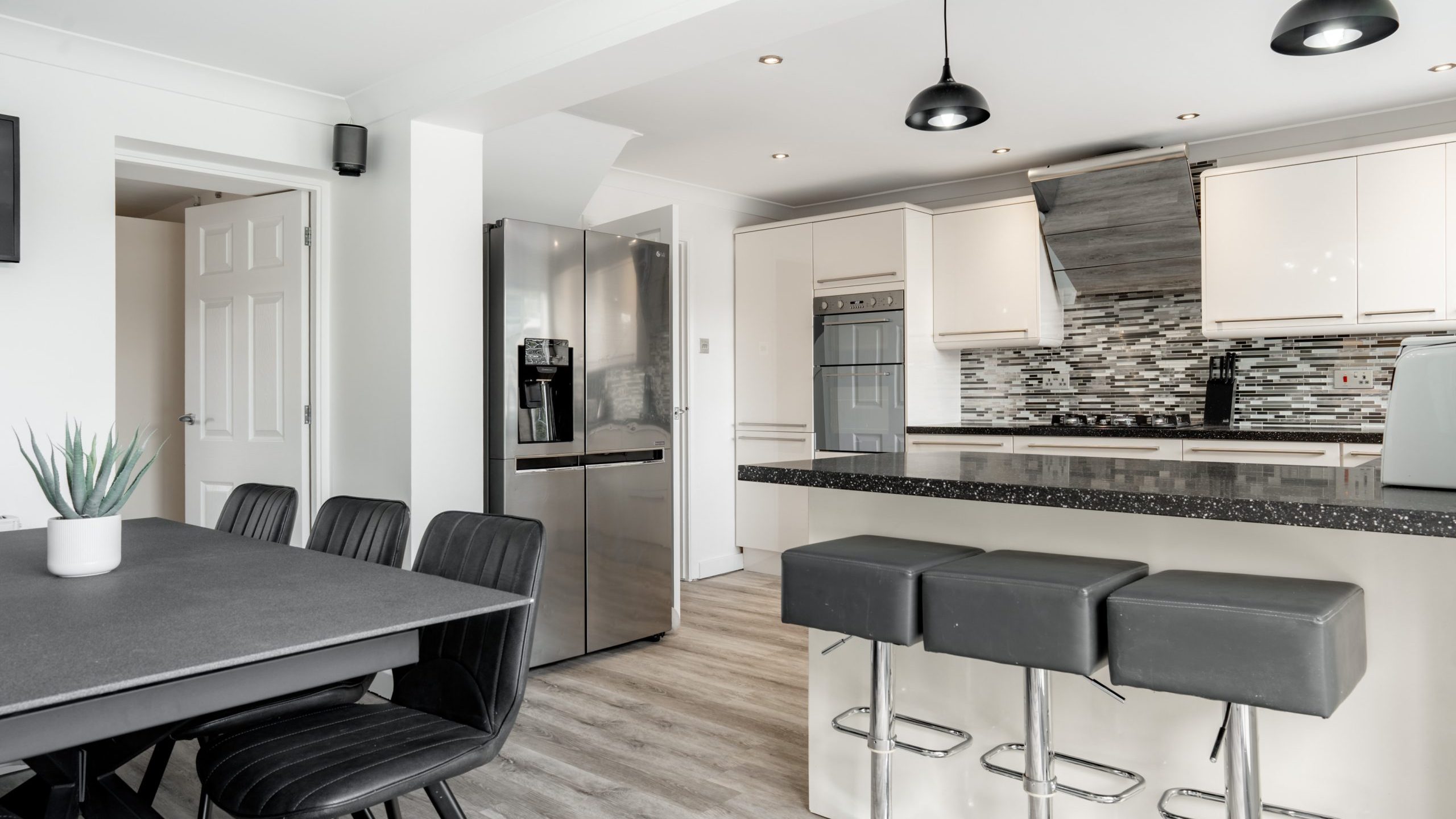 Modern kitchen with a dining area, featuring black chairs, a gray table, bar stools, stainless steel appliances, white cabinets, a tiled backsplash, and pendant lights. A small potted plant decorates the table.