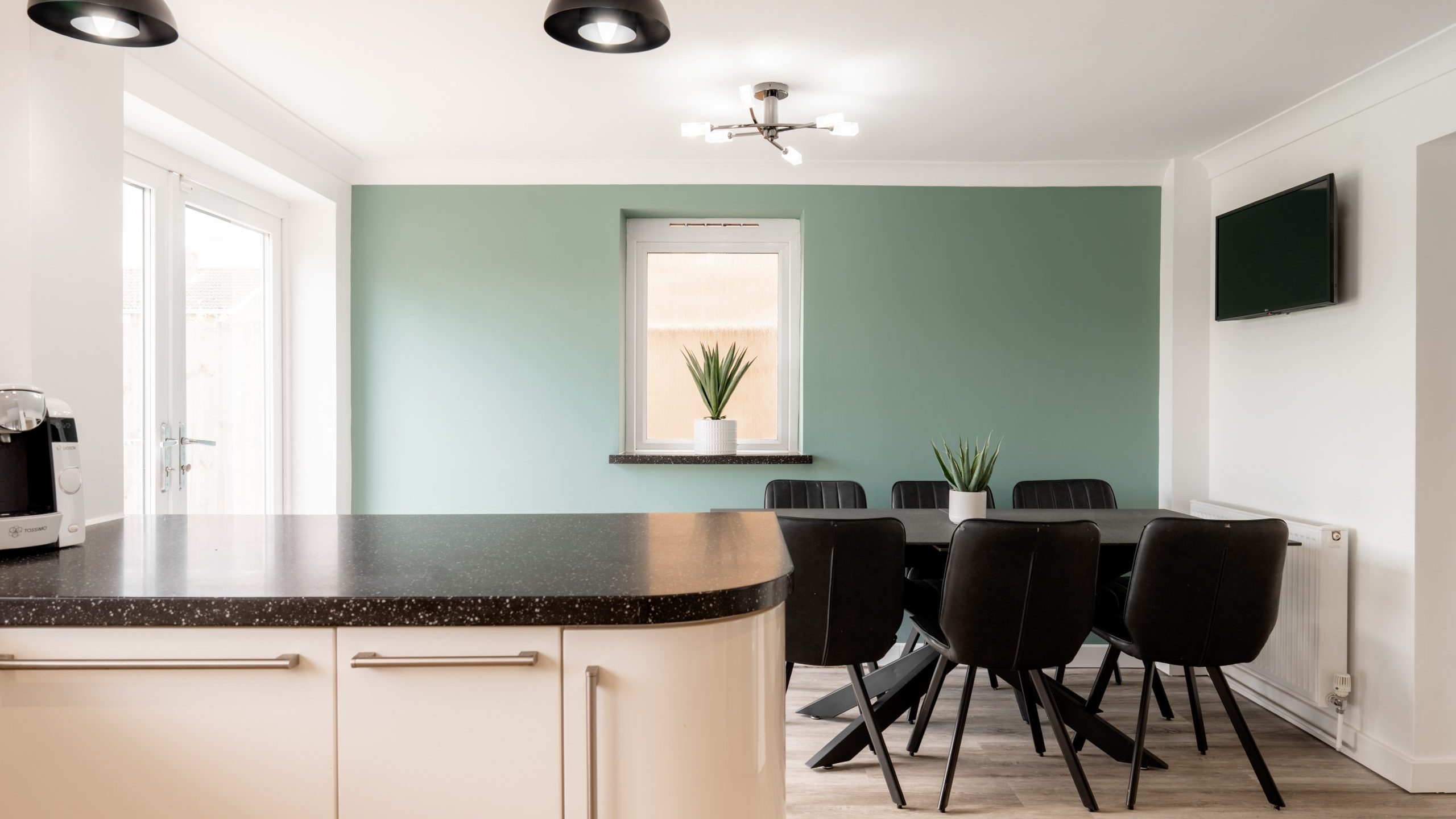 Modern kitchen and dining area with black chairs around a round table, green accent wall with a window, potted plants, overhead lights, and a wall-mounted TV. Light wood flooring and cream-colored cabinets are visible.