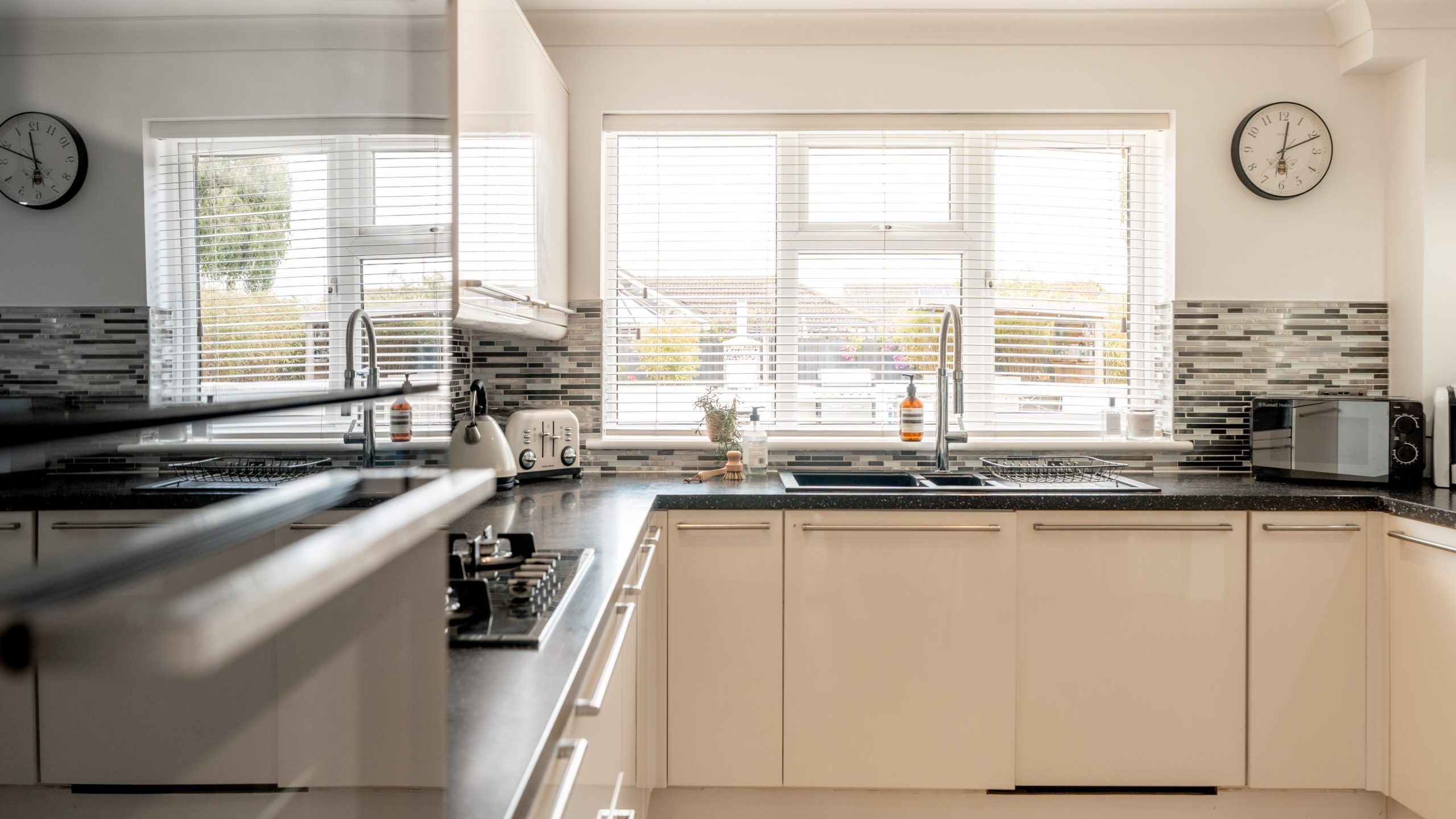 Bright modern kitchen with white cabinets, black countertops, stainless steel appliances, a toaster, kettle, microwave, and a window letting in natural light. Two clocks and tile backsplash decorate the walls.