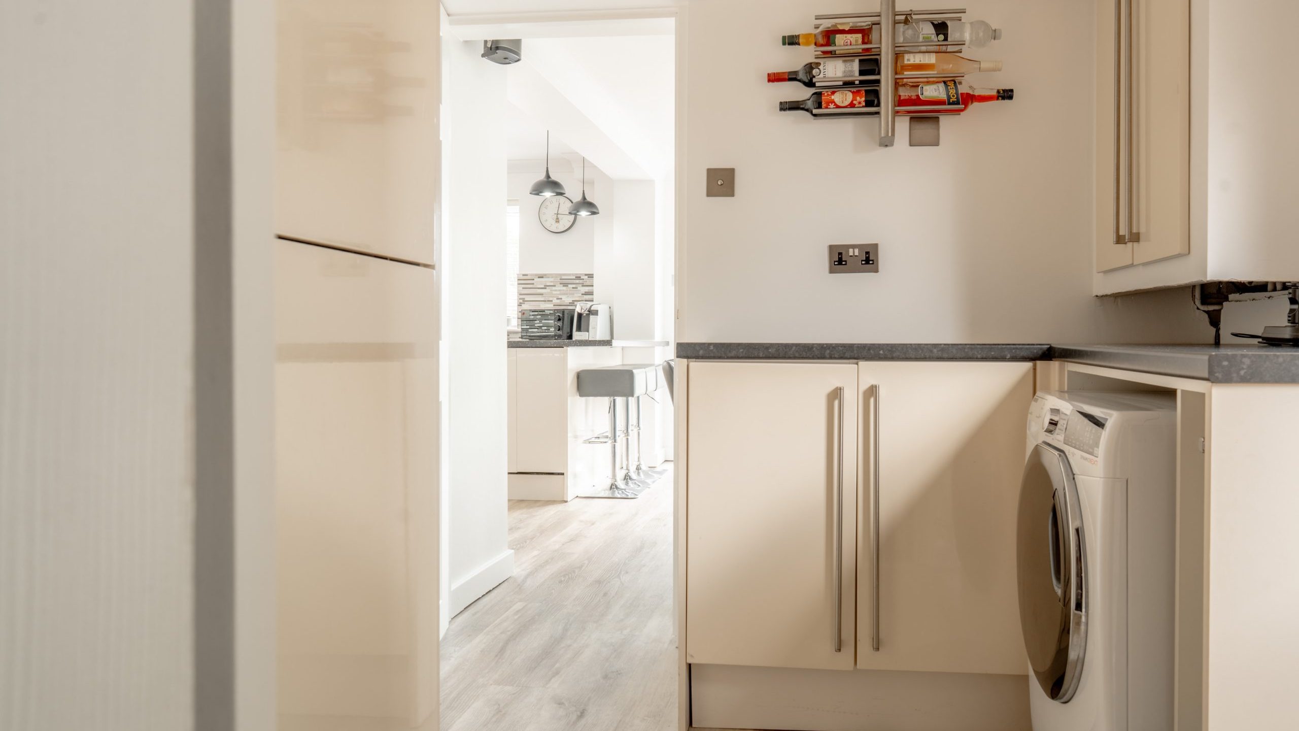 Modern kitchen with light-colored cabinets, a washing machine, and a wine rack on the wall. The space has light wood flooring and leads to a bright dining area with a clock and bar stools in the background.