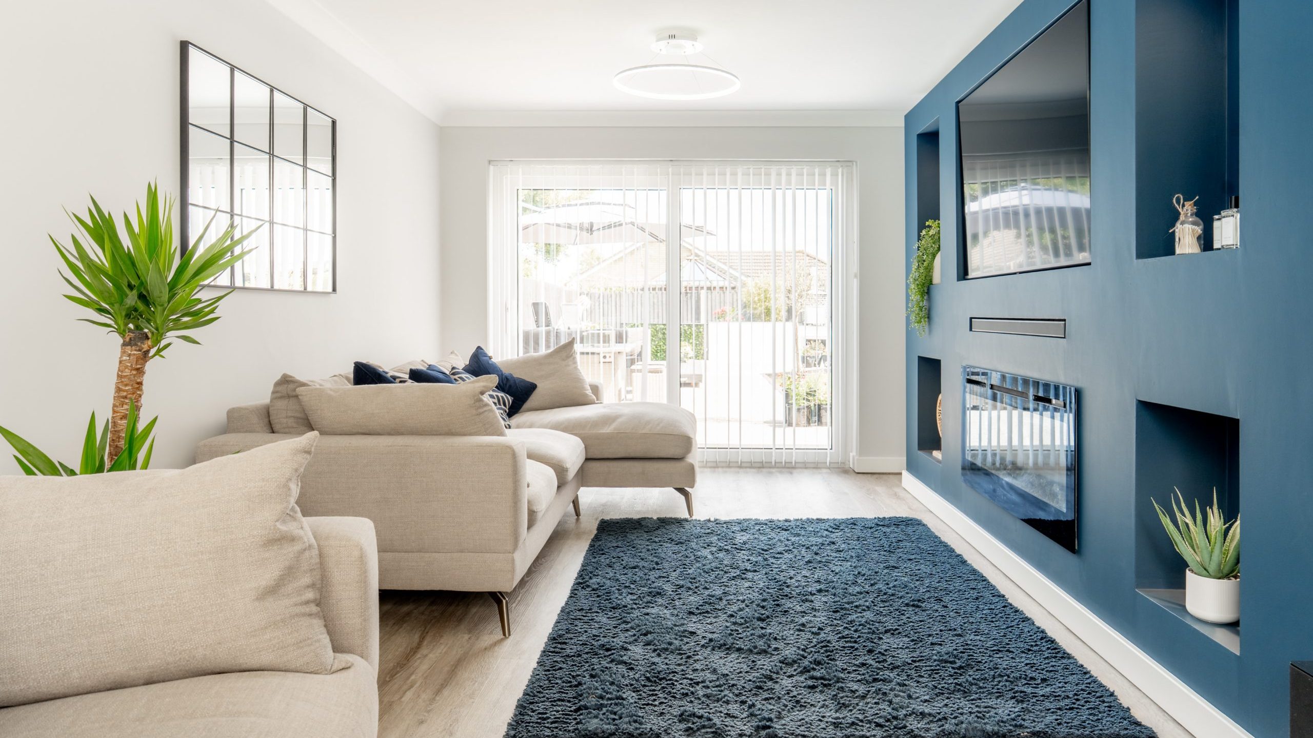 Modern living room with light beige sofas, a blue accent wall with built-in TV and shelves, a blue rug, potted plants, and large glass doors letting in natural light from outside.