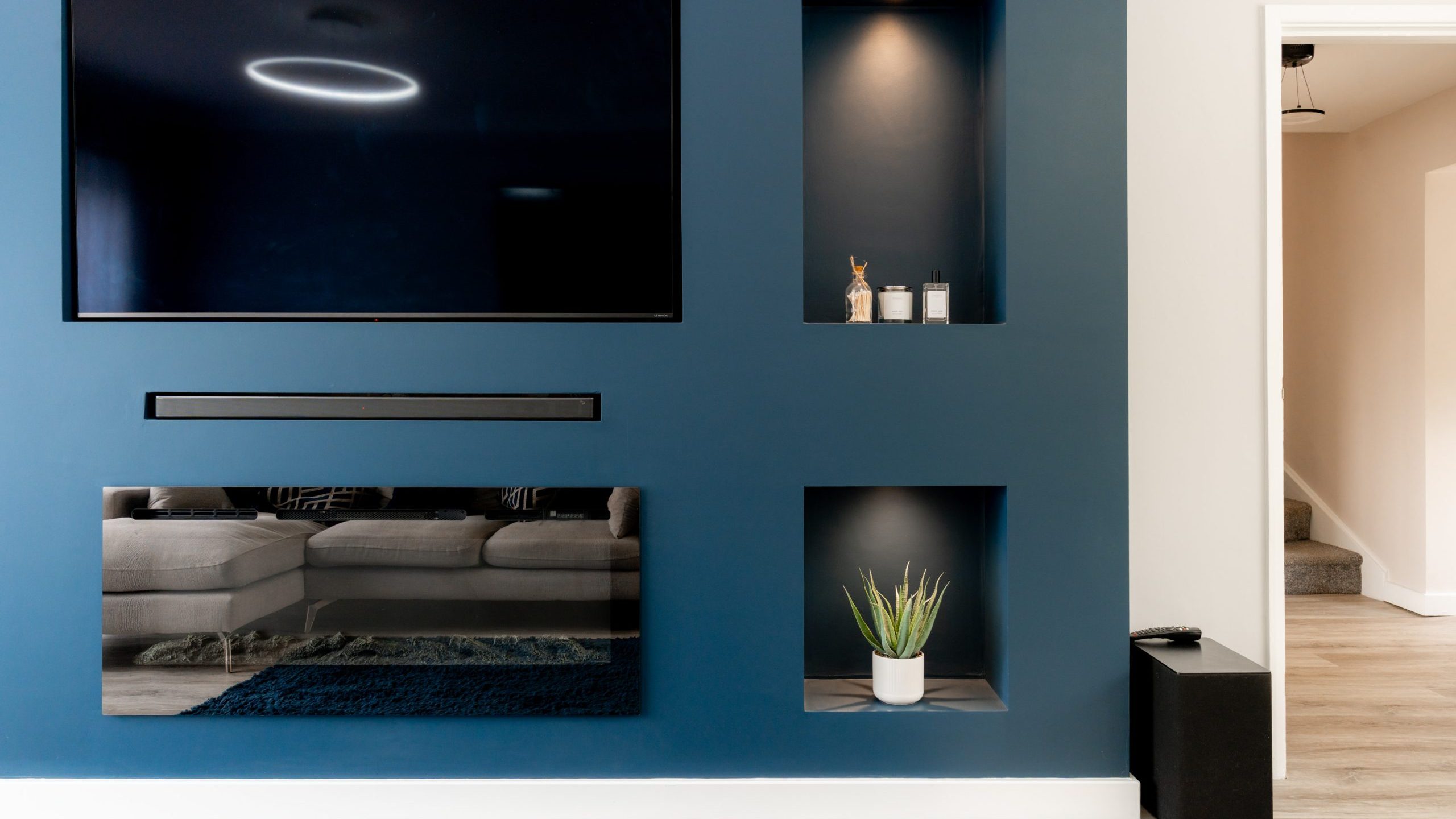 A modern living room wall with a mounted TV, black soundbar, and shelving. The blue wall has two built-in niches displaying small decor and a potted plant. A hallway and stairs are visible to the right.