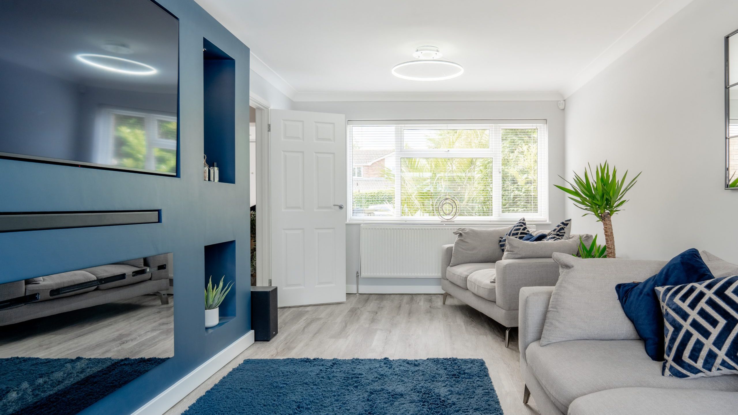 A modern living room with two light gray sofas, blue and white cushions, a blue accent wall with a flat-screen TV, a blue rug, light wood flooring, large window, and indoor plants. Bright and airy atmosphere.
