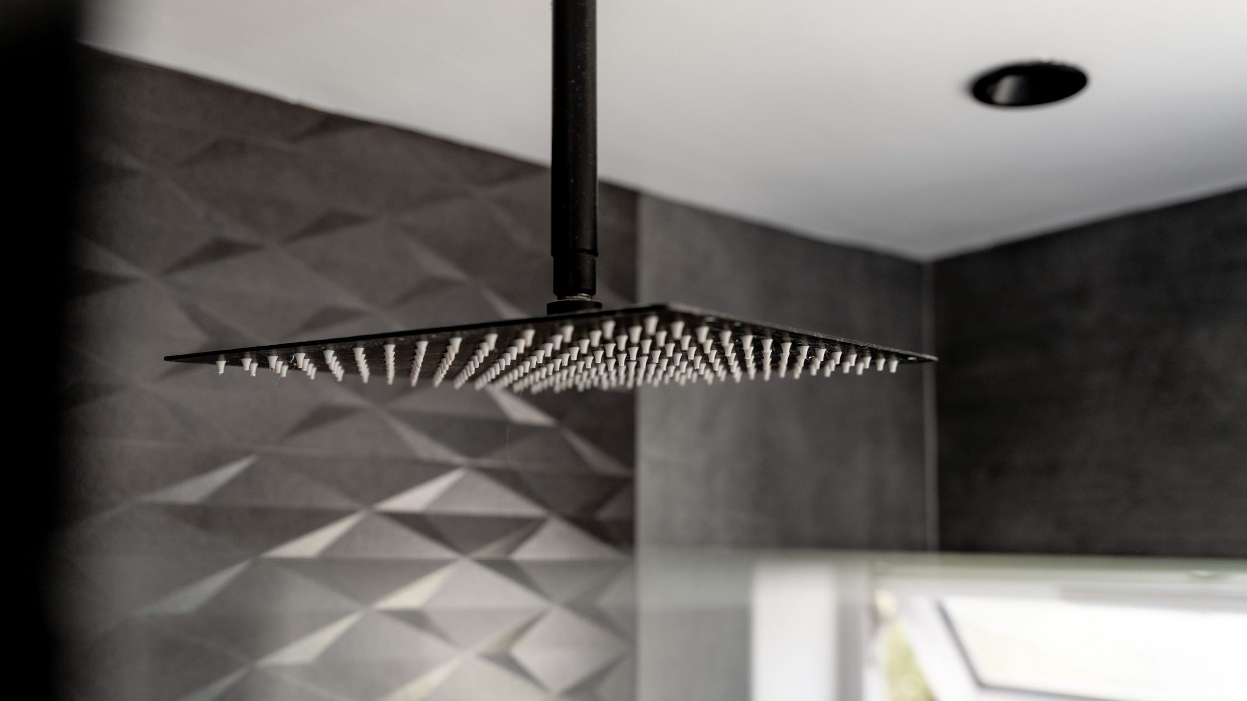 A close-up of a modern, square, overhead showerhead in a bathroom with dark, textured, geometric-patterned walls and a small window letting in natural light.