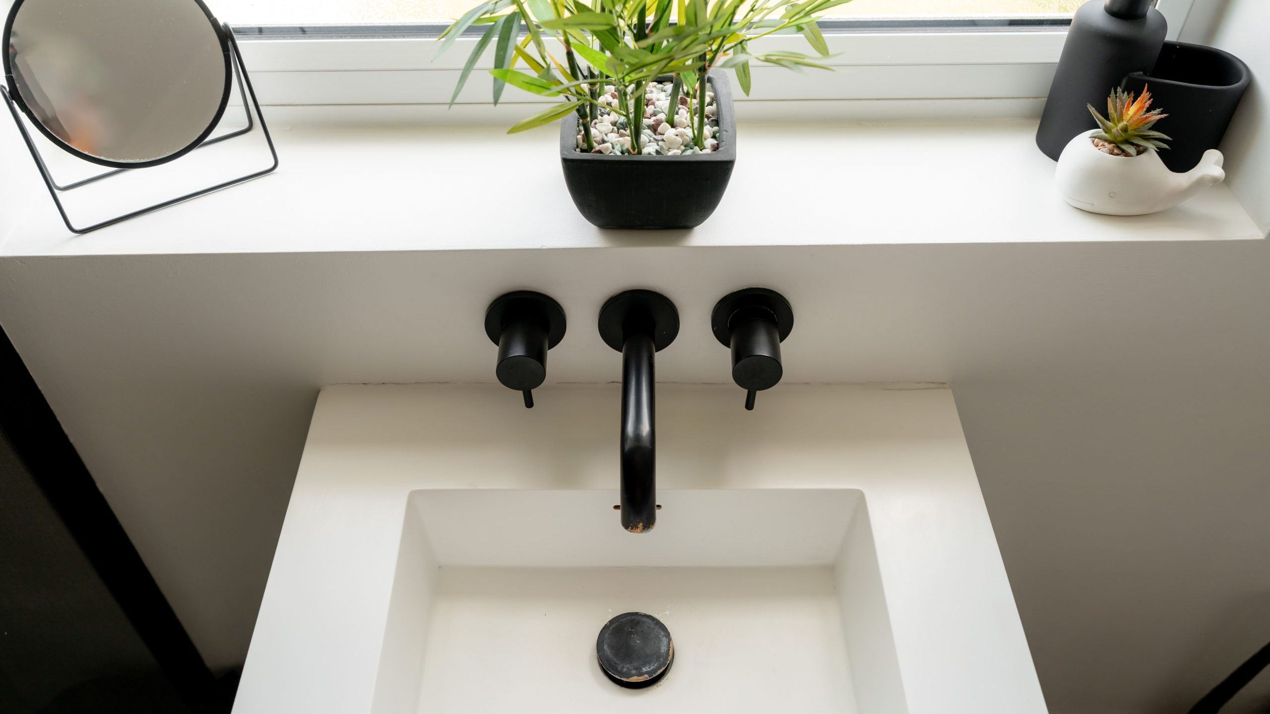 Modern white bathroom sink with a matte black faucet and handles, a small potted plant, and a round mirror on the windowsill above. A soap dispenser and a decorative item are also present.