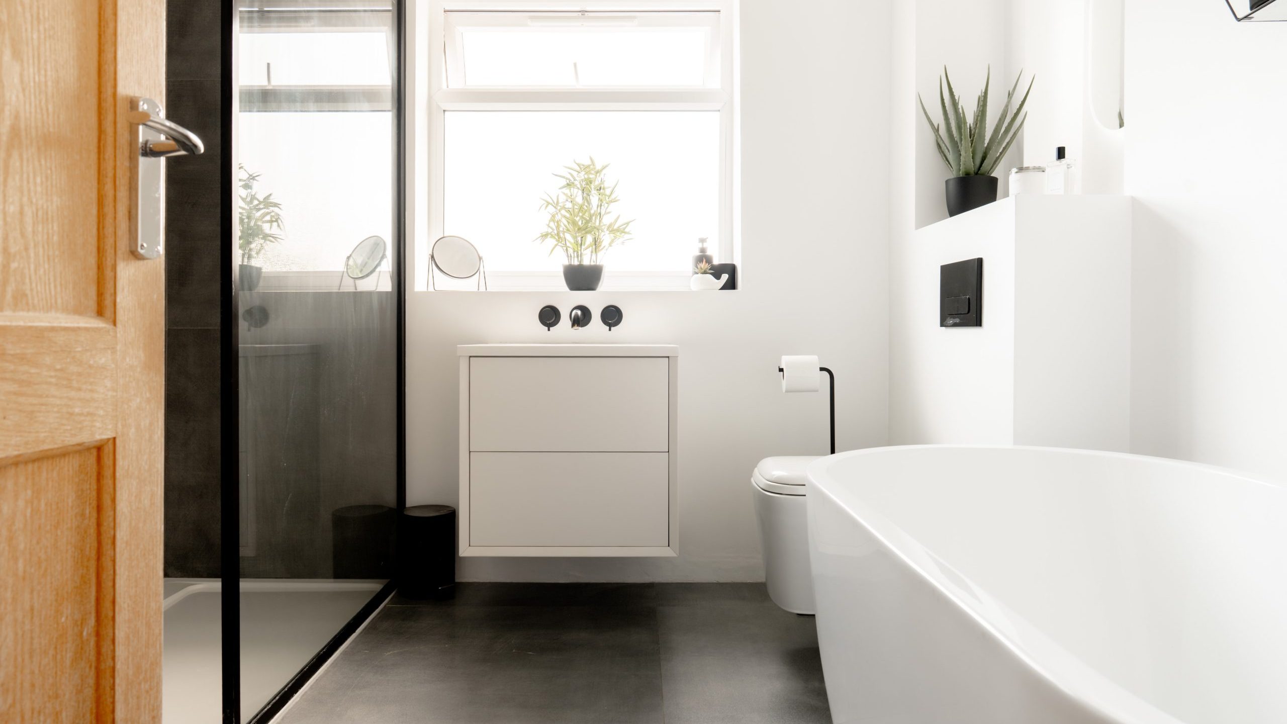 Modern bathroom with a freestanding bathtub, glass-enclosed shower, floating vanity, white walls, gray floor tiles, potted plants on the windowsill, and minimalist decor. Natural light comes through the window.