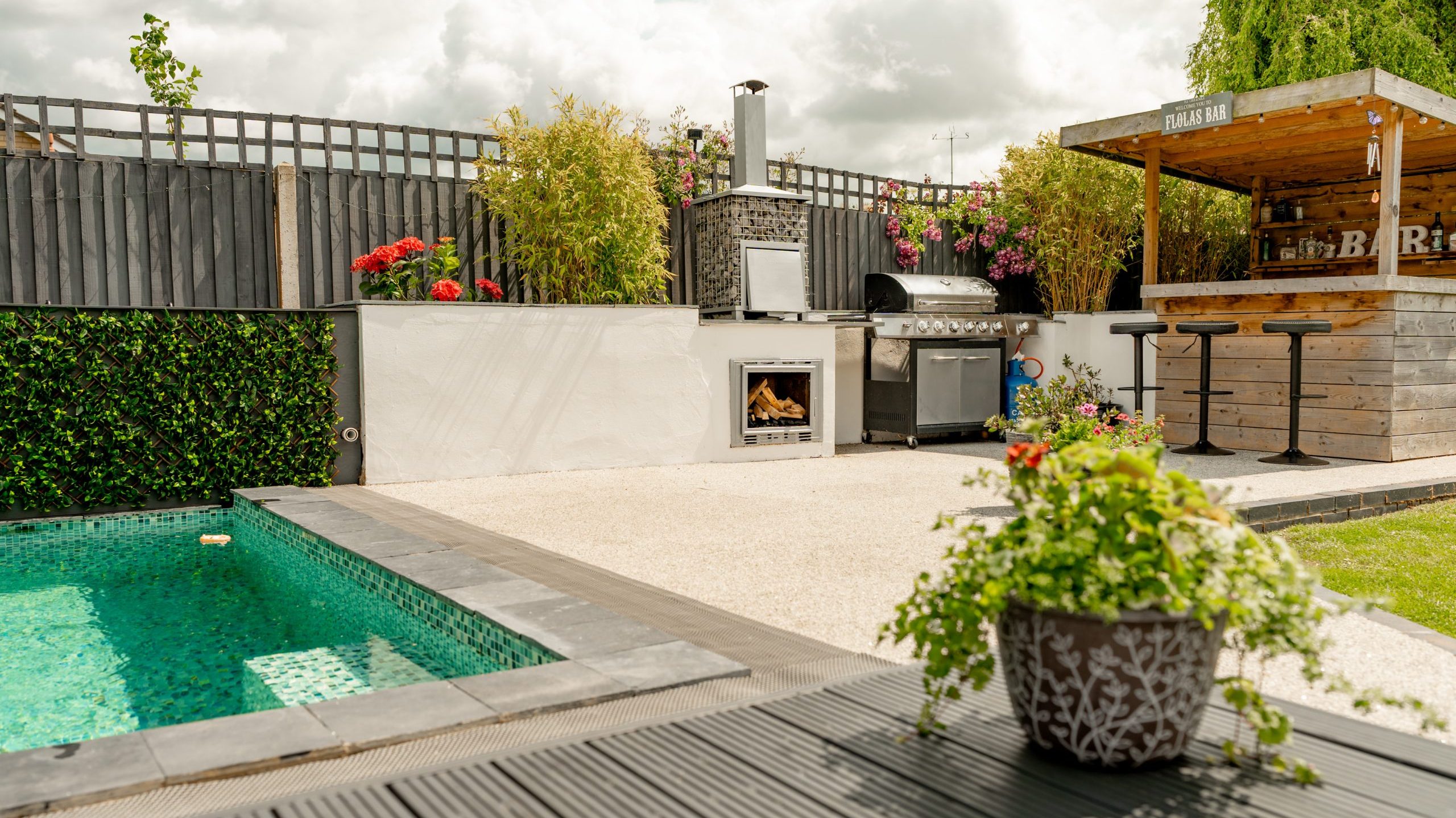 Modern backyard with a swimming pool, outdoor kitchen with a pizza oven and grill, a bar area with stools, potted plants, and a wooden privacy fence under a partly cloudy sky.
