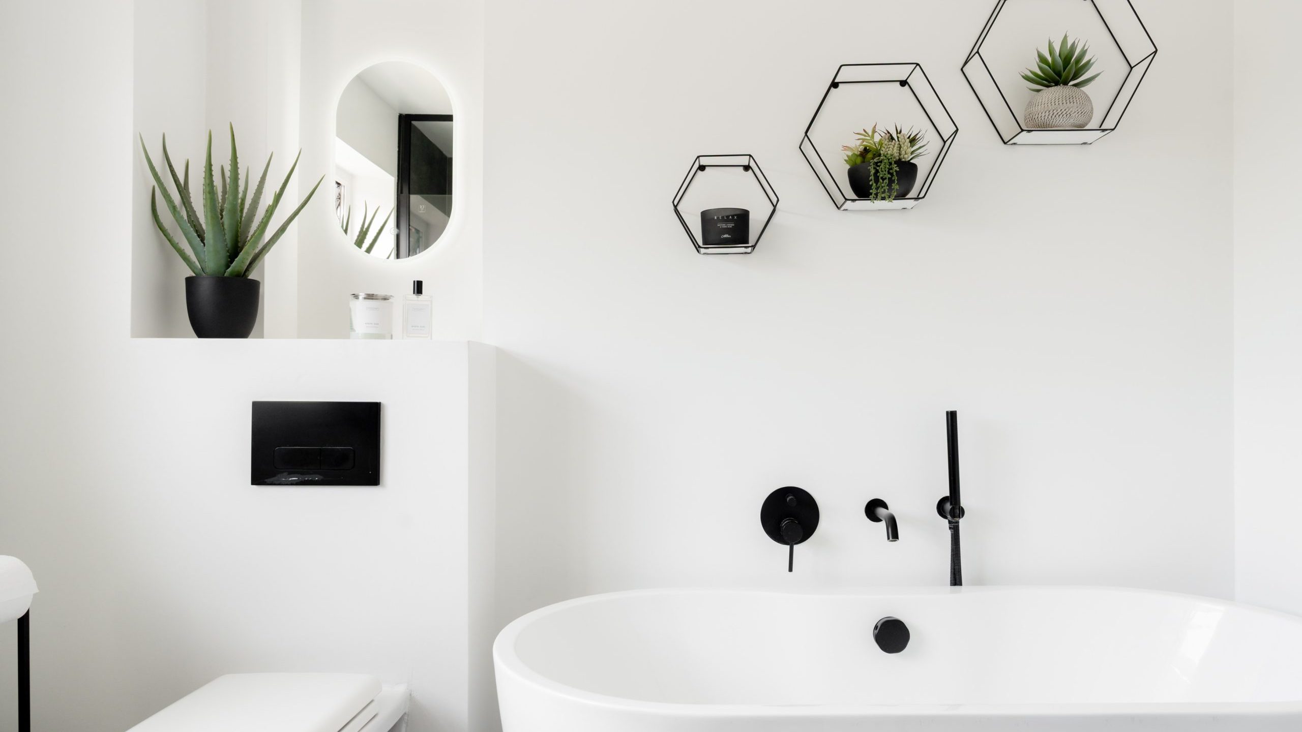 Minimalist white bathroom with a modern oval bathtub, black wall-mounted fixtures, a toilet, potted plants, and hexagonal wall shelves holding small plants and decor items. A round mirror hangs above a shelf.