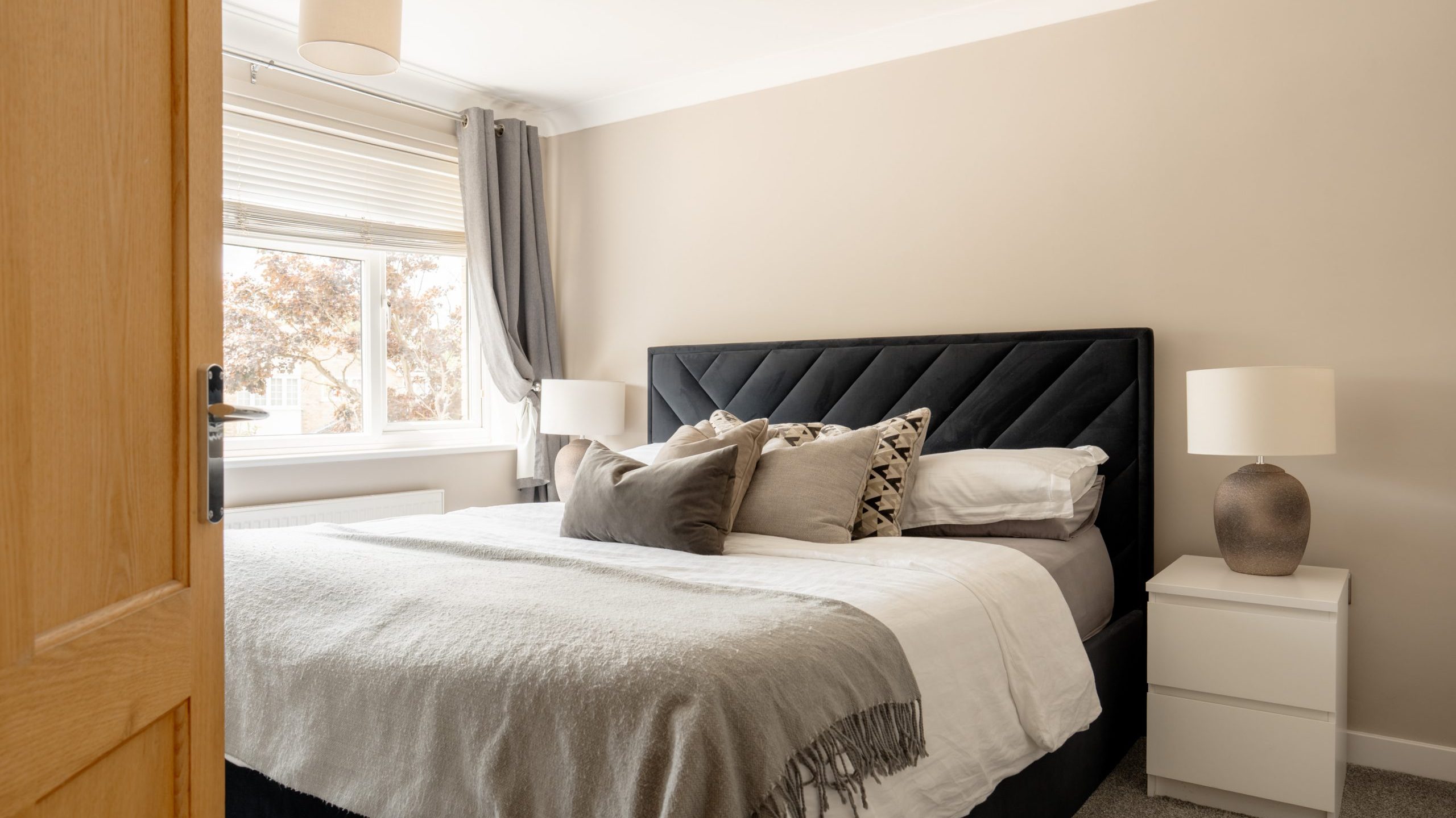 A modern bedroom with a large bed featuring neutral-toned bedding and decorative pillows, two white nightstands with lamps, a window with light curtains, and beige walls creating a cozy atmosphere.