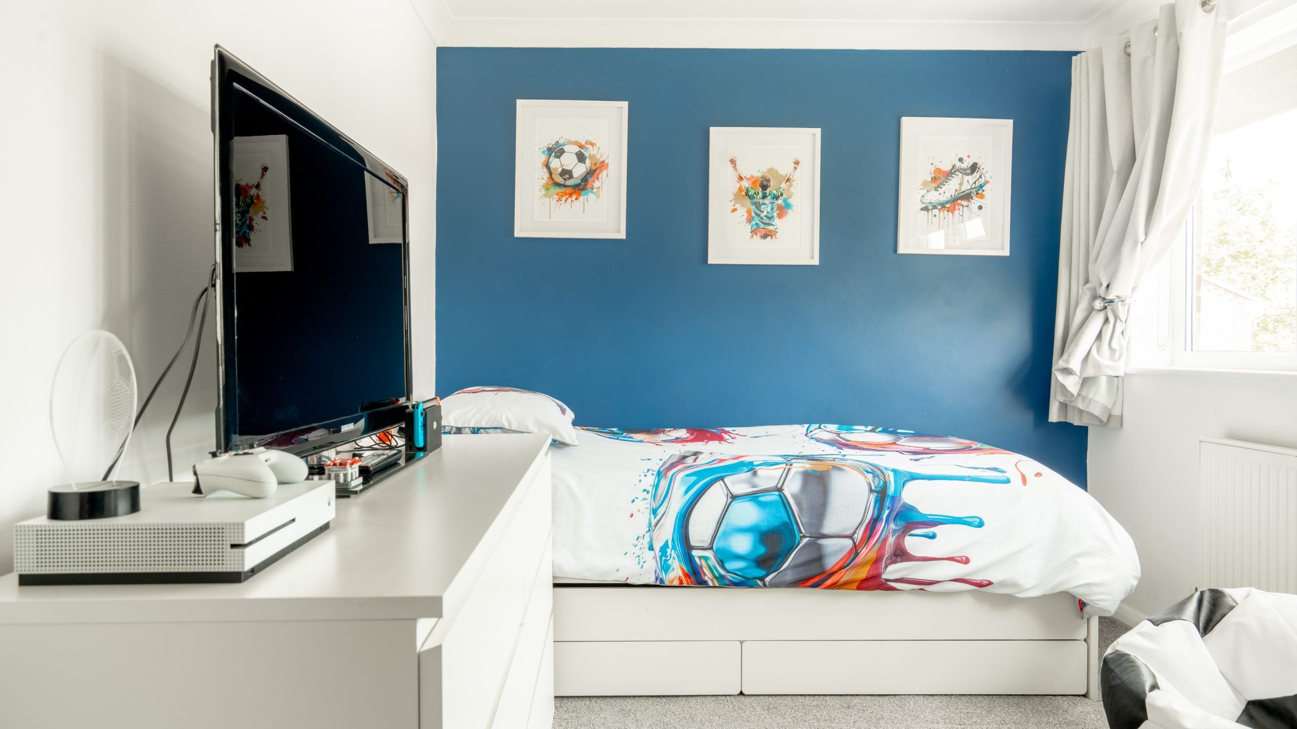 A modern bedroom with a single bed featuring soccer-themed bedding, a blue accent wall with three framed prints, a white dresser with a TV and gaming console, and a window with light curtains.