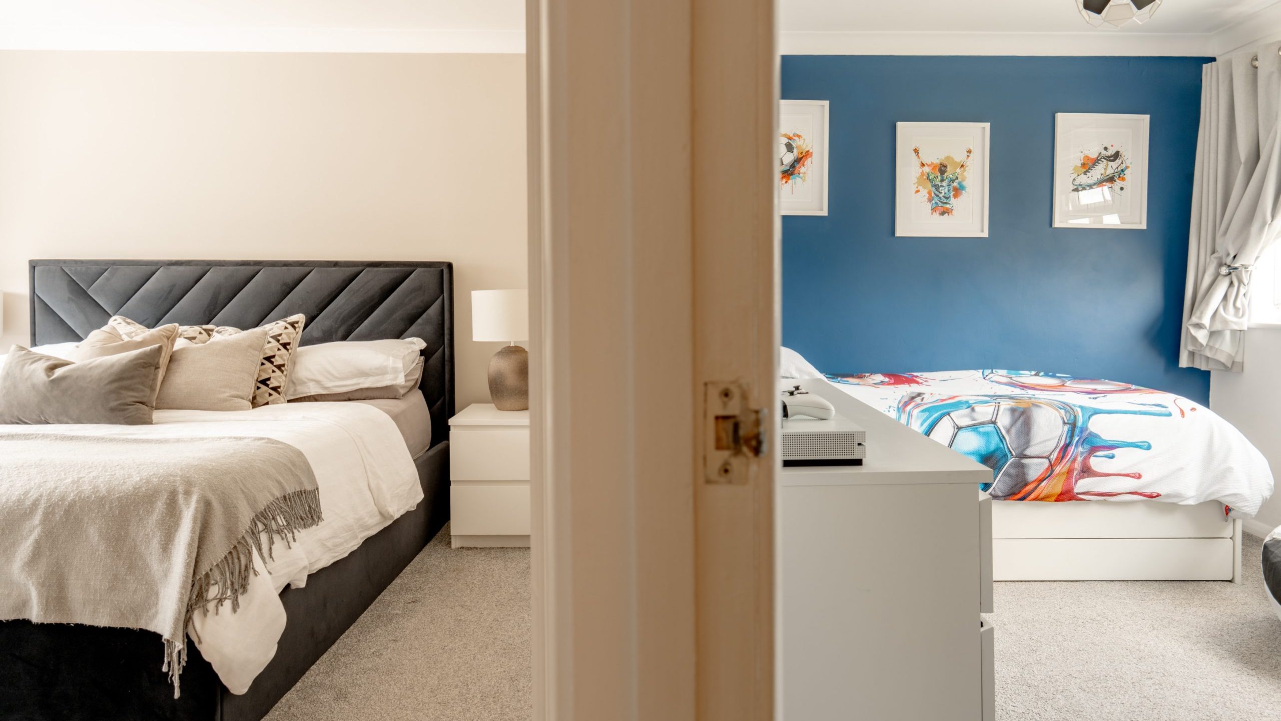 A view through an open doorway showing two bedrooms; one with a dark upholstered bed and neutral decor, the other with a blue accent wall, colorful bedding, and framed artwork above the bed.