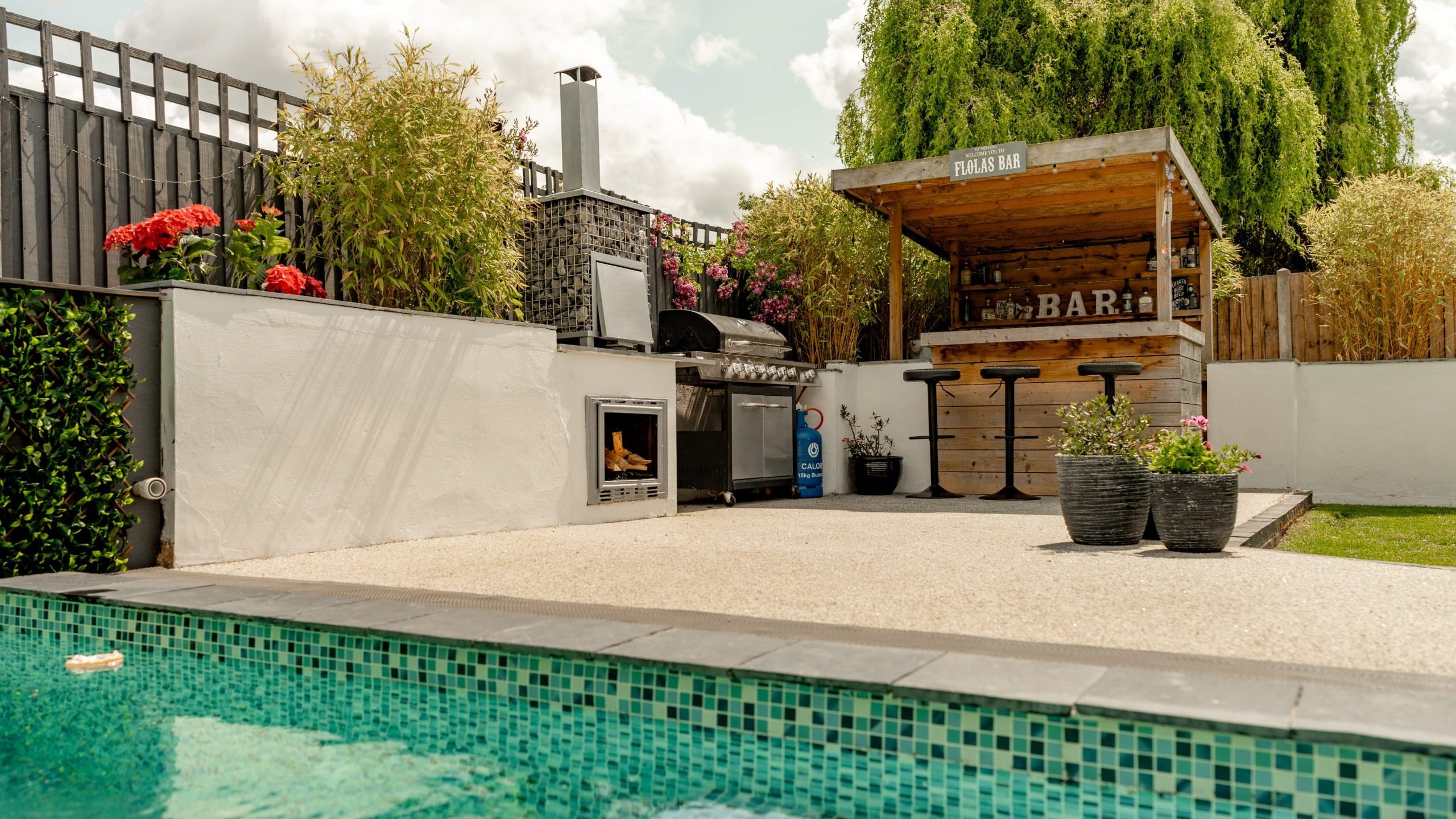 A backyard patio with a small wooden bar, barbecue grill, and fireplace, surrounded by plants and flowers, next to a turquoise-tiled swimming pool under a partly cloudy sky.