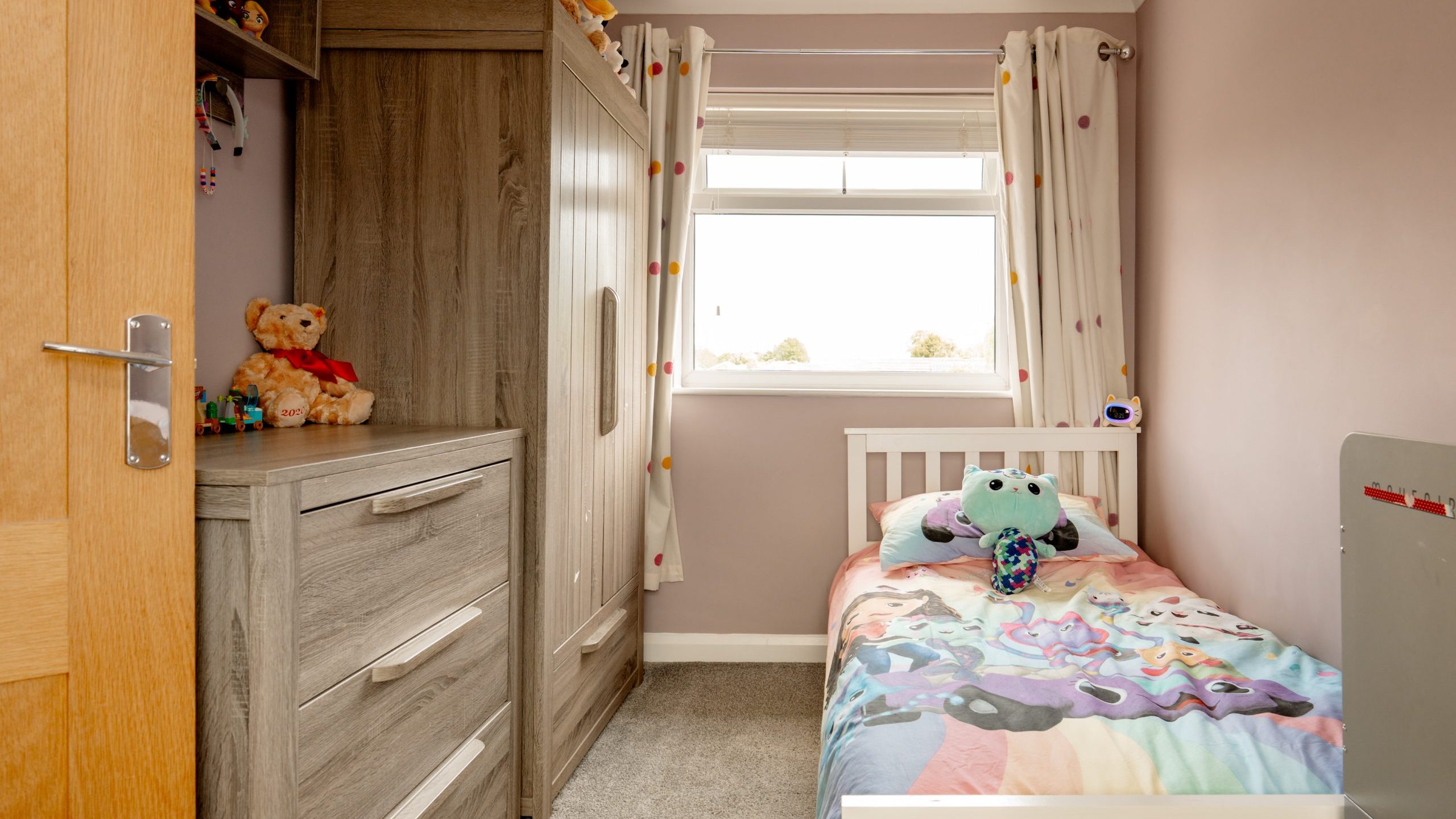 A small, neatly organized child’s bedroom with a single bed covered in a colorful blanket, a large wardrobe, plush toys on shelves, light brown walls, and a window with white curtains letting in natural light.