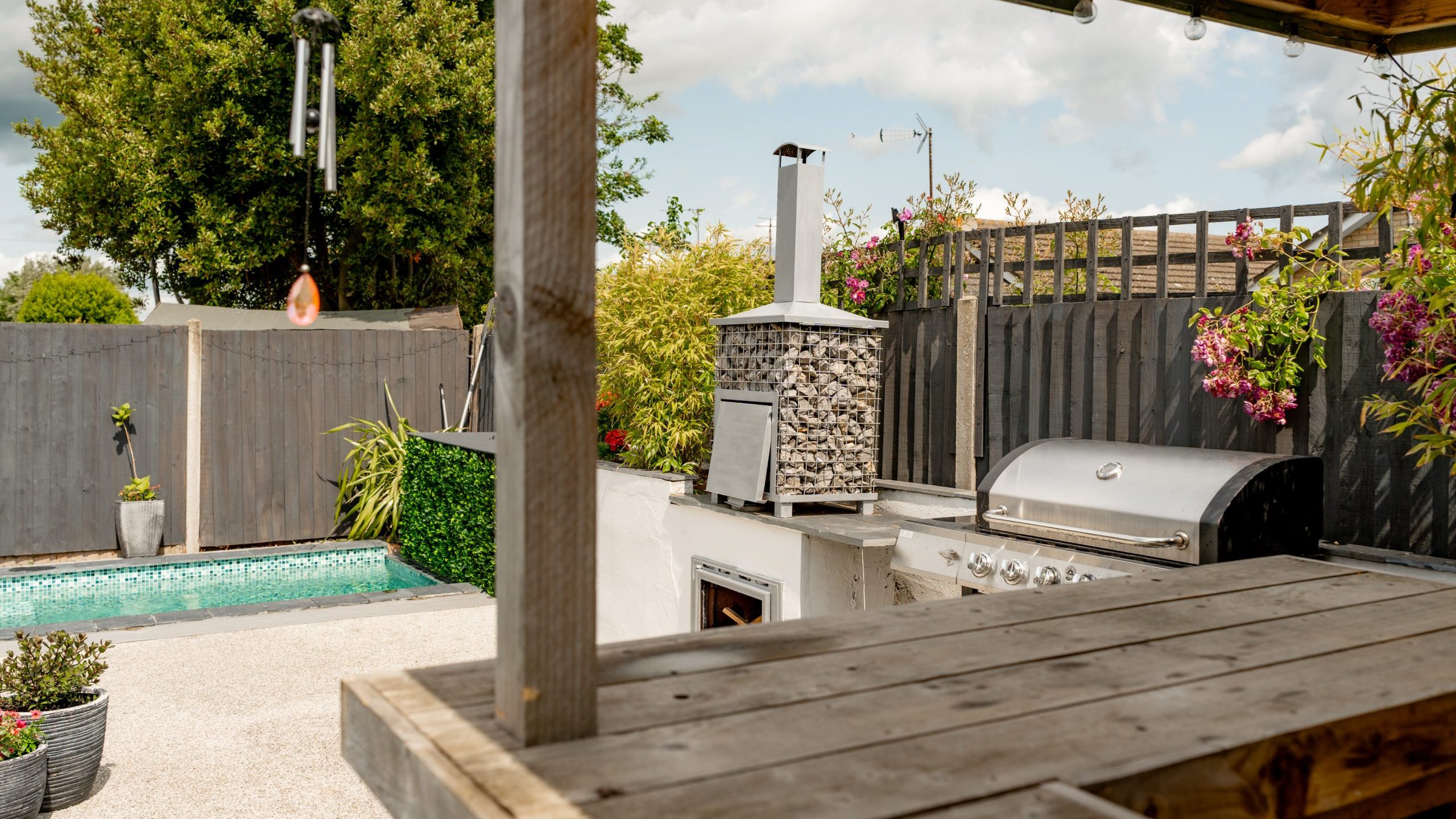 Outdoor patio scene with a wooden counter in the foreground, a grill, and a pizza oven. There's a small swimming pool, potted plants, a fence, and a tree under a partly cloudy sky.