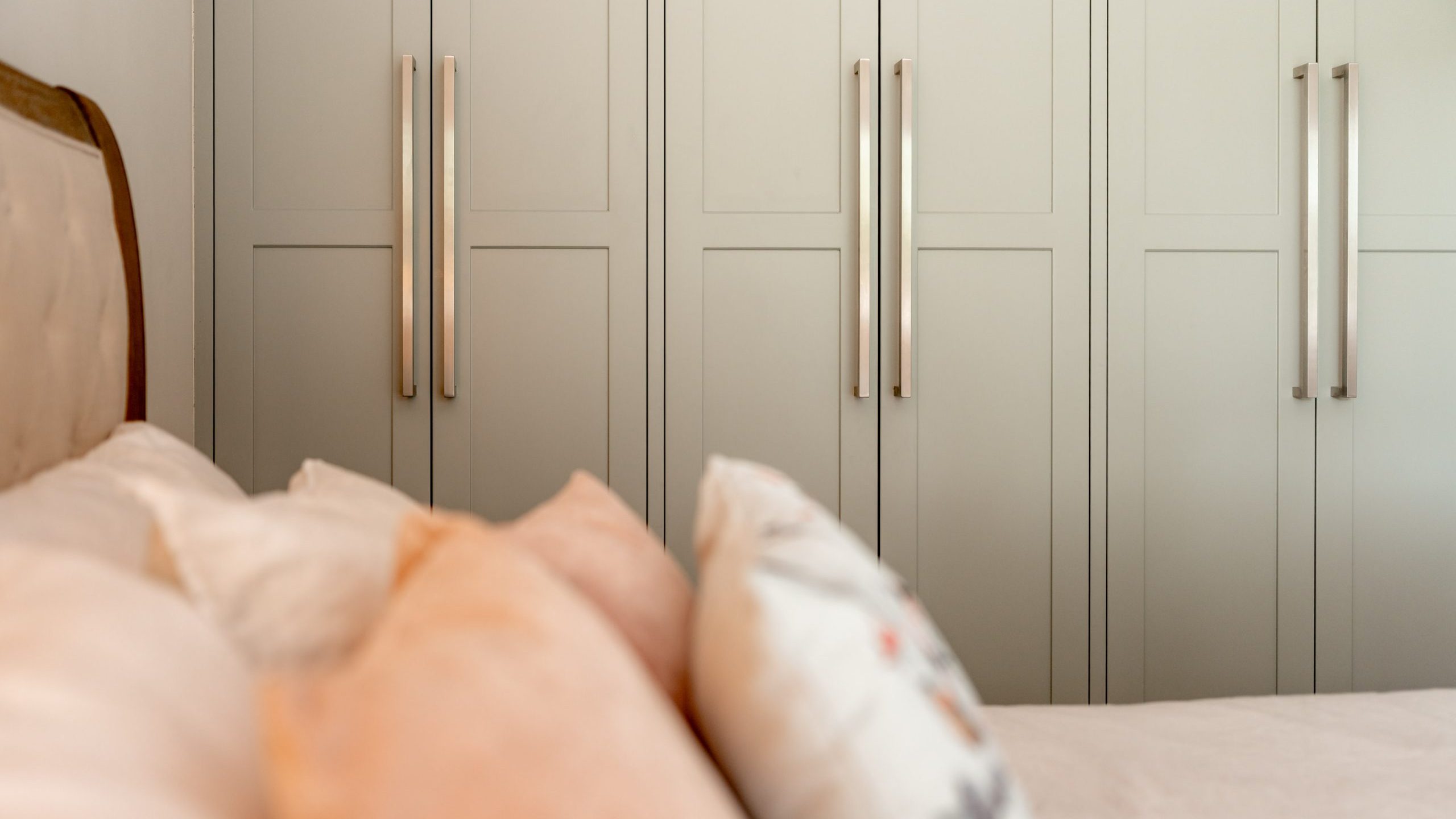 A close-up of a bed with pastel-colored pillows in front of a wall of tall, light-colored wardrobes with long, metallic handles. The scene appears calm and tidy, suggesting a cozy bedroom.