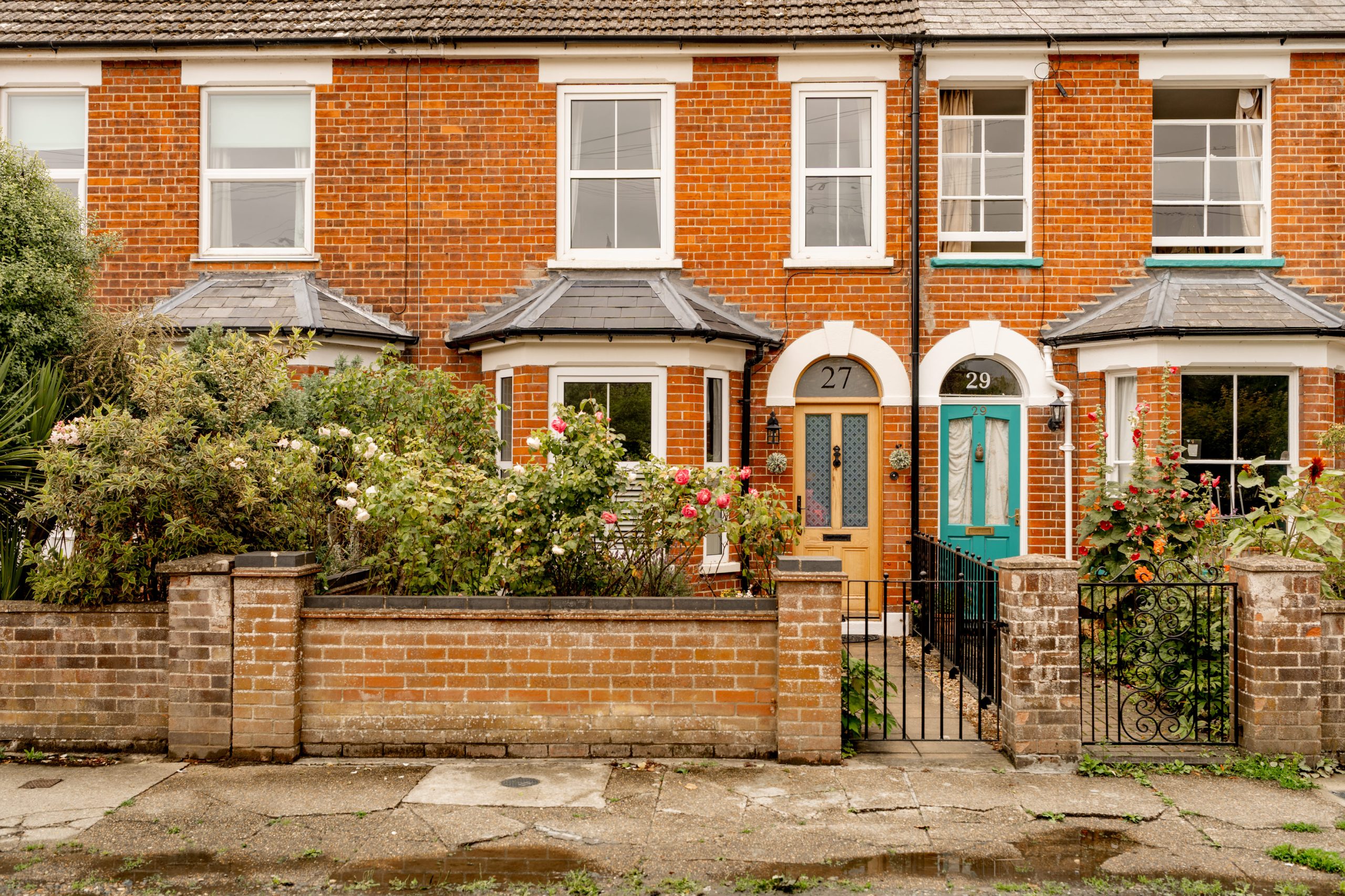 Two adjoining red-brick terraced houses with bay windows, small front gardens with flowering shrubs, and arched doorways. Both houses have stone steps and brick or wrought iron fences along the sidewalk.