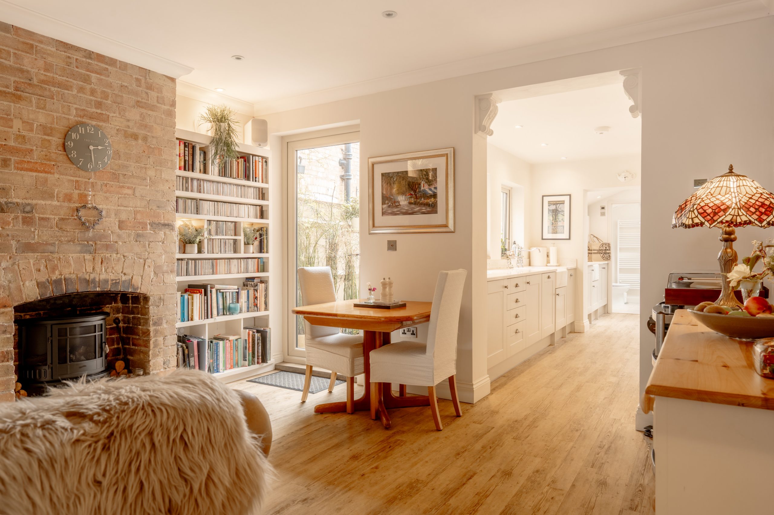 Bright, cozy dining area with a wooden table and two white chairs near a brick fireplace, bookshelves, and a kitchen with white cabinets in the background. Warm lighting and homey decor create an inviting atmosphere.