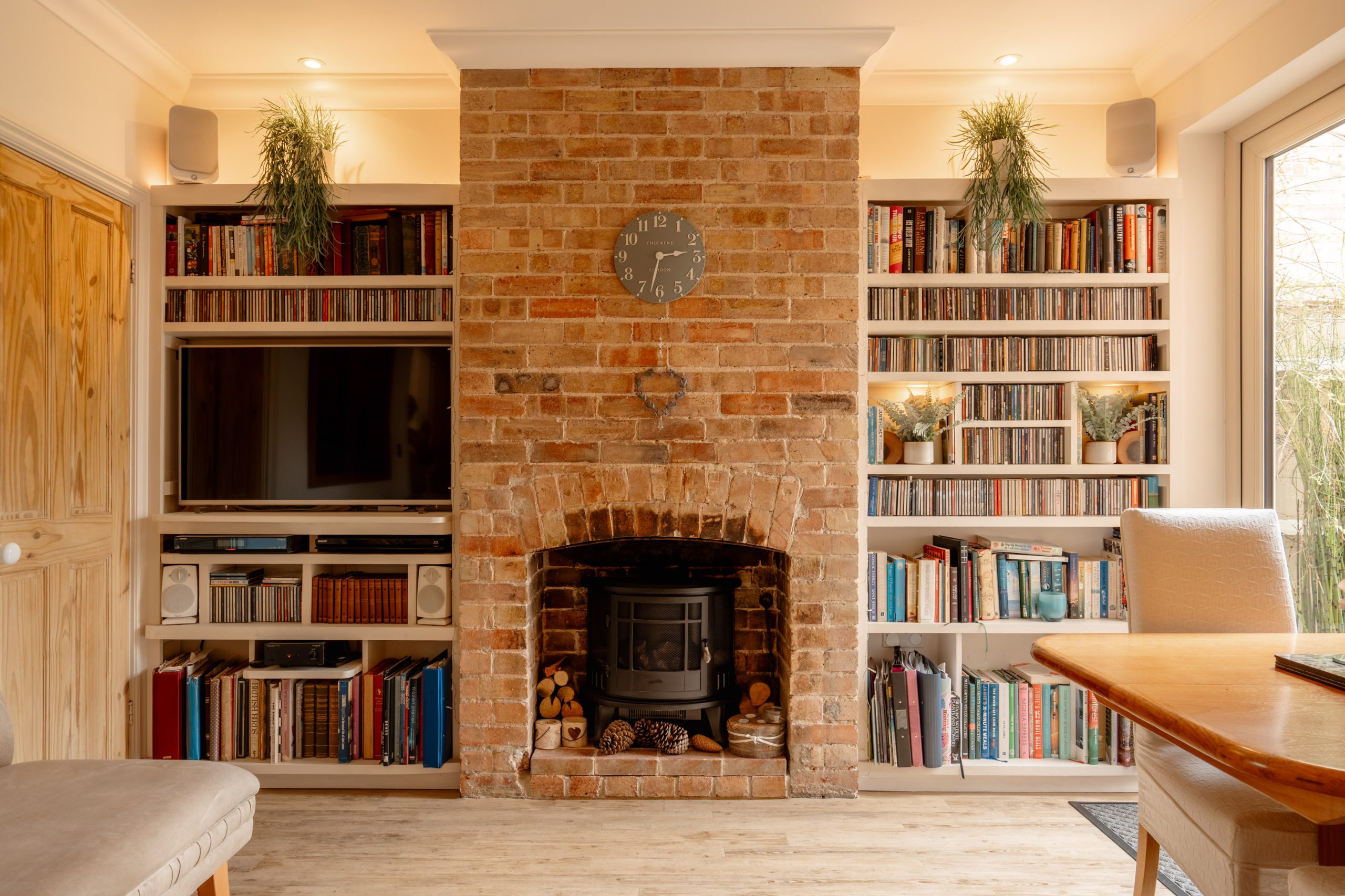 A cozy living room with a brick fireplace at the center, flanked by bookshelves filled with books and a TV on the left. Plants sit on top, and a clock hangs above the fireplace. Warm, inviting lighting brightens the space.