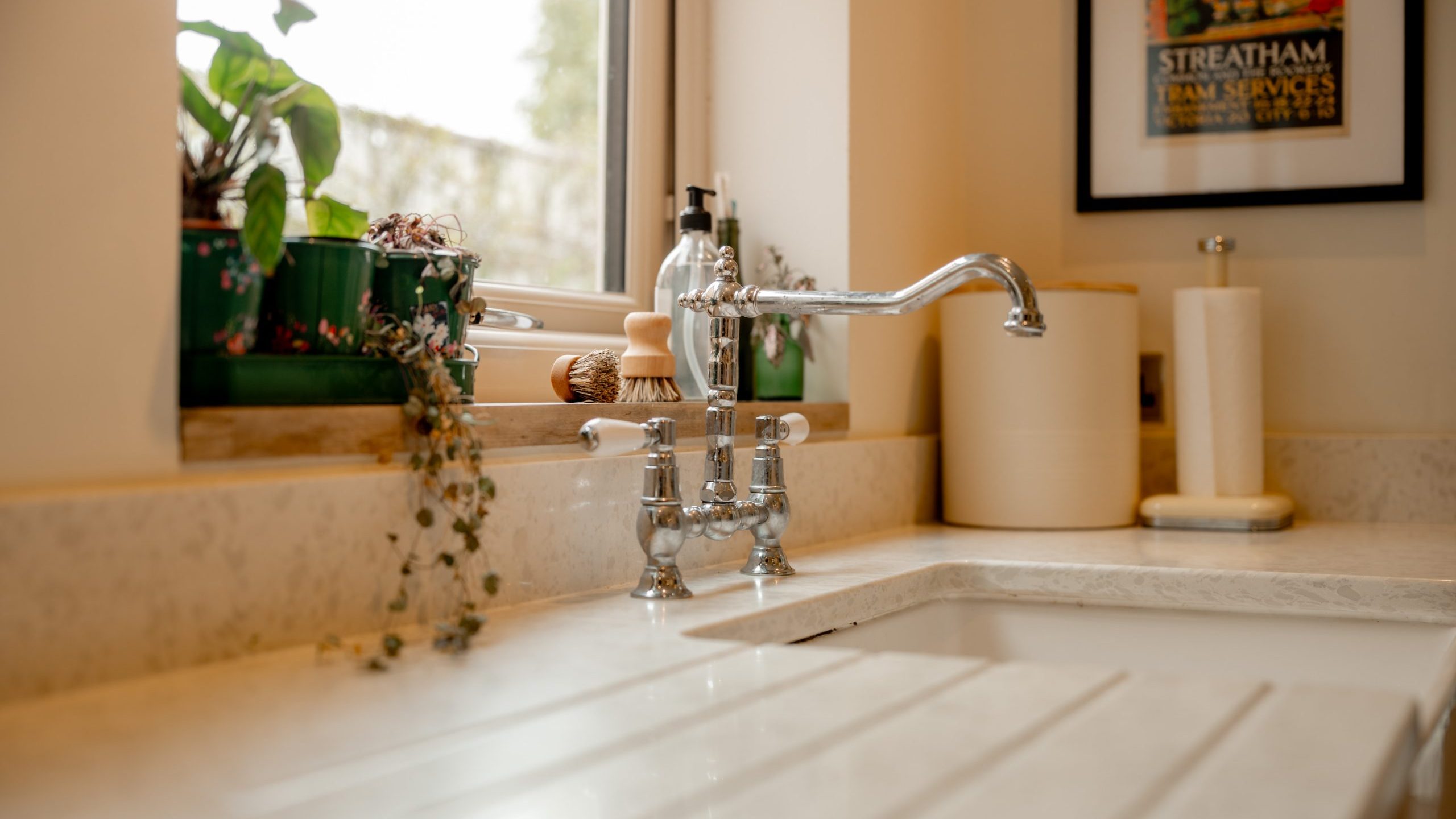 A clean, modern kitchen sink with a shiny chrome faucet, houseplants on the windowsill, a roll of paper towels, and a framed picture on the wall. Natural light comes in through the window.