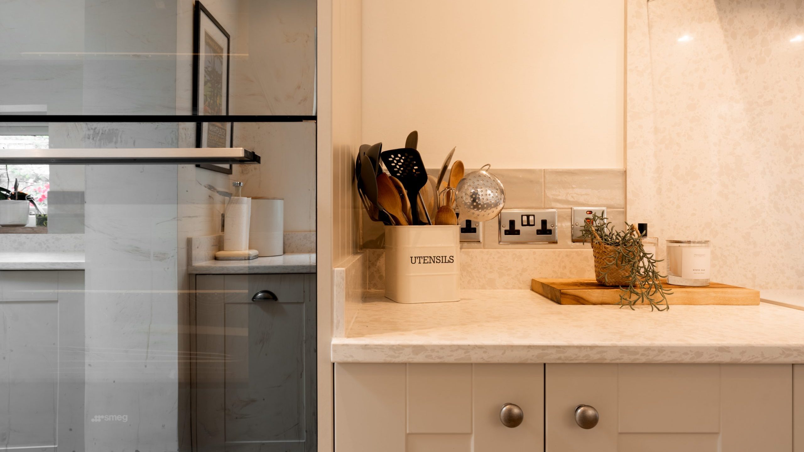 Modern kitchen countertop with a beige utensil holder containing wooden spoons, a cutting board with herbs, and an oven or microwave built into the cabinetry on the left. The surface appears clean and organized.