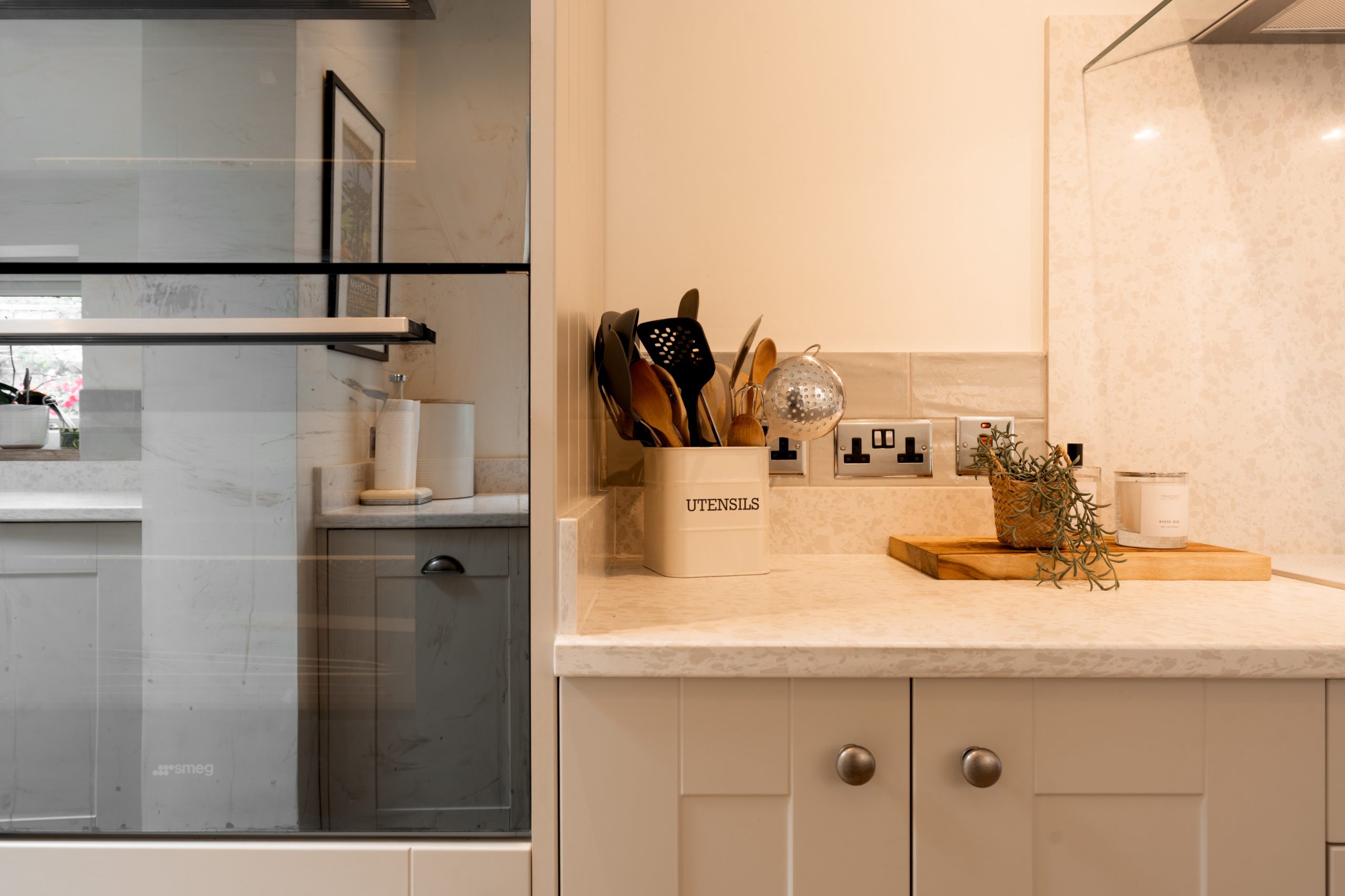 Modern kitchen countertop with a beige utensil holder containing wooden spoons, a cutting board with herbs, and an oven or microwave built into the cabinetry on the left. The surface appears clean and organized.