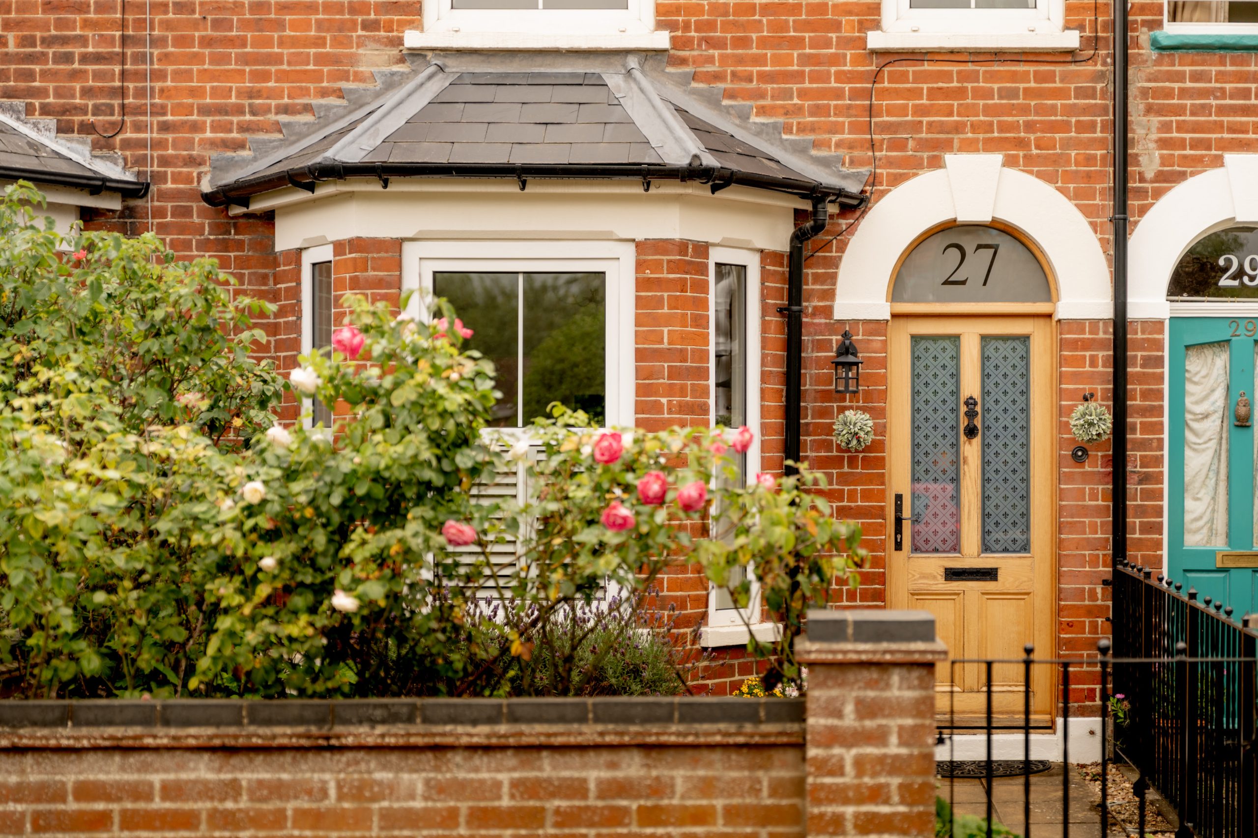 A red-brick house with a bay window, wooden front door numbered 27, white trim, and climbing pink roses in the front garden behind a low brick wall.