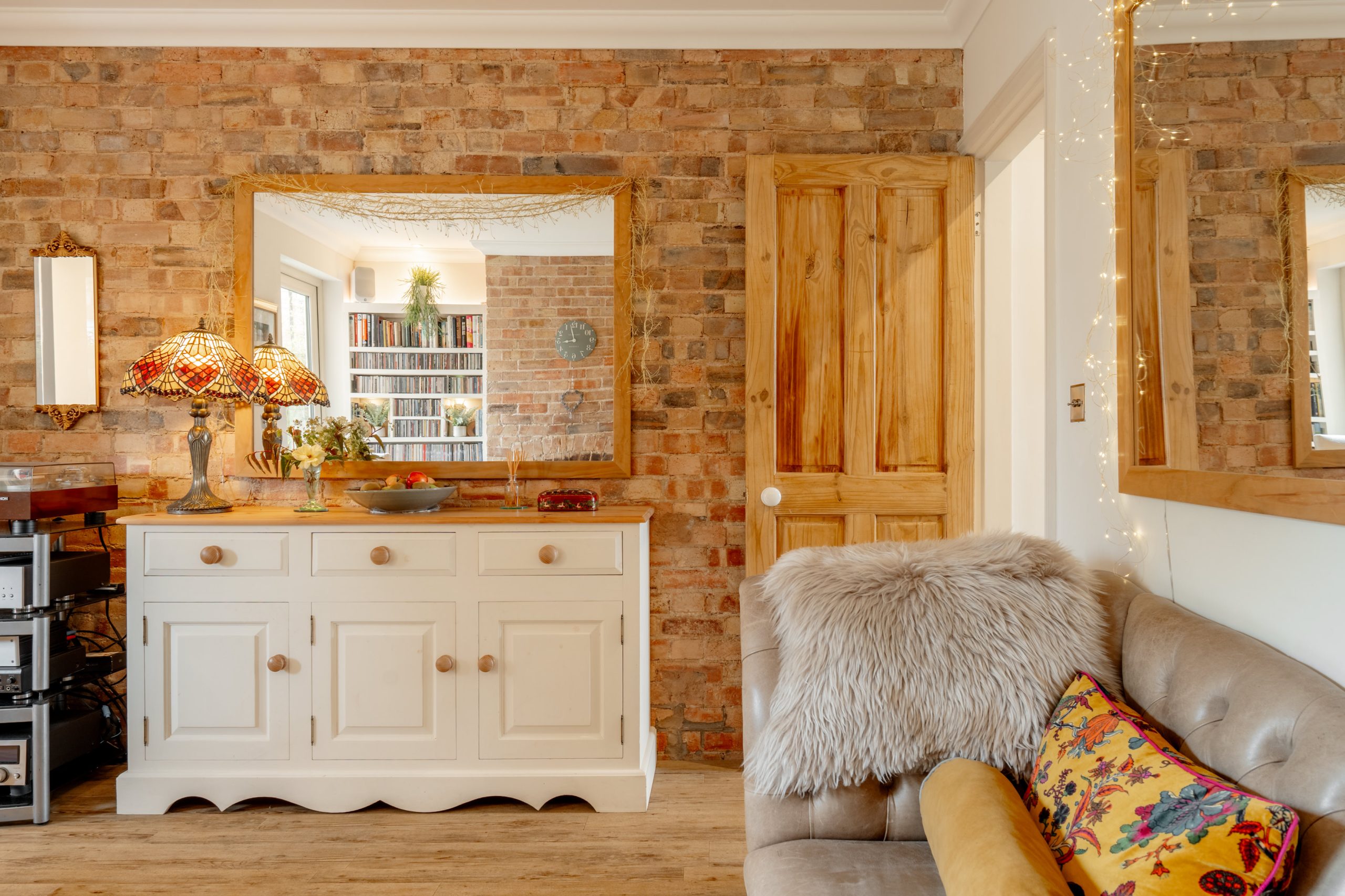 Cozy living room with exposed brick walls, a large mirror, a white sideboard with colorful lamps, a wooden door, a bookshelf reflected in the mirror, and a beige sofa with a fluffy throw and floral pillow.