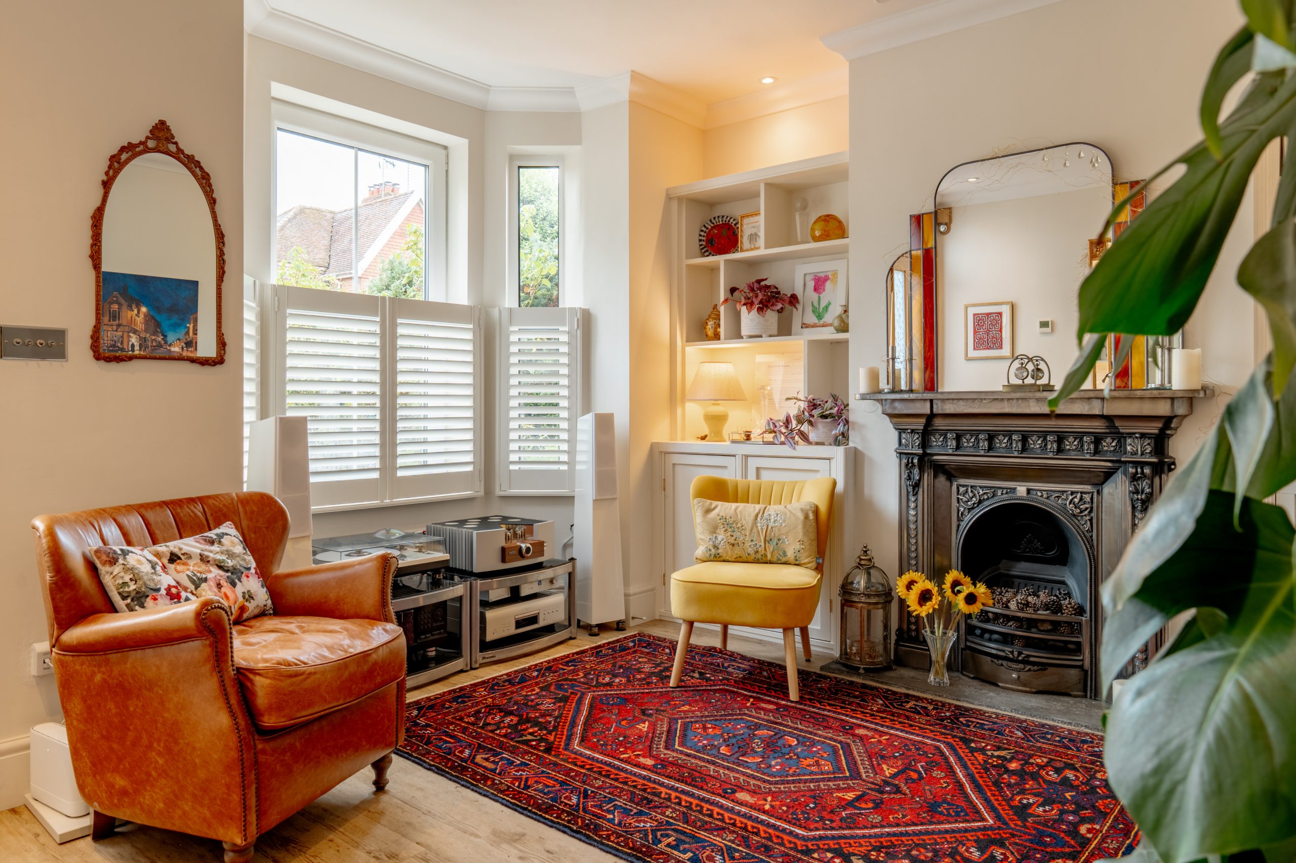Cozy living room with a leather armchair, yellow chair, ornate fireplace, decorative mirror, red patterned rug, indoor plants, and shelves with books and decor near a bright window with shutters.