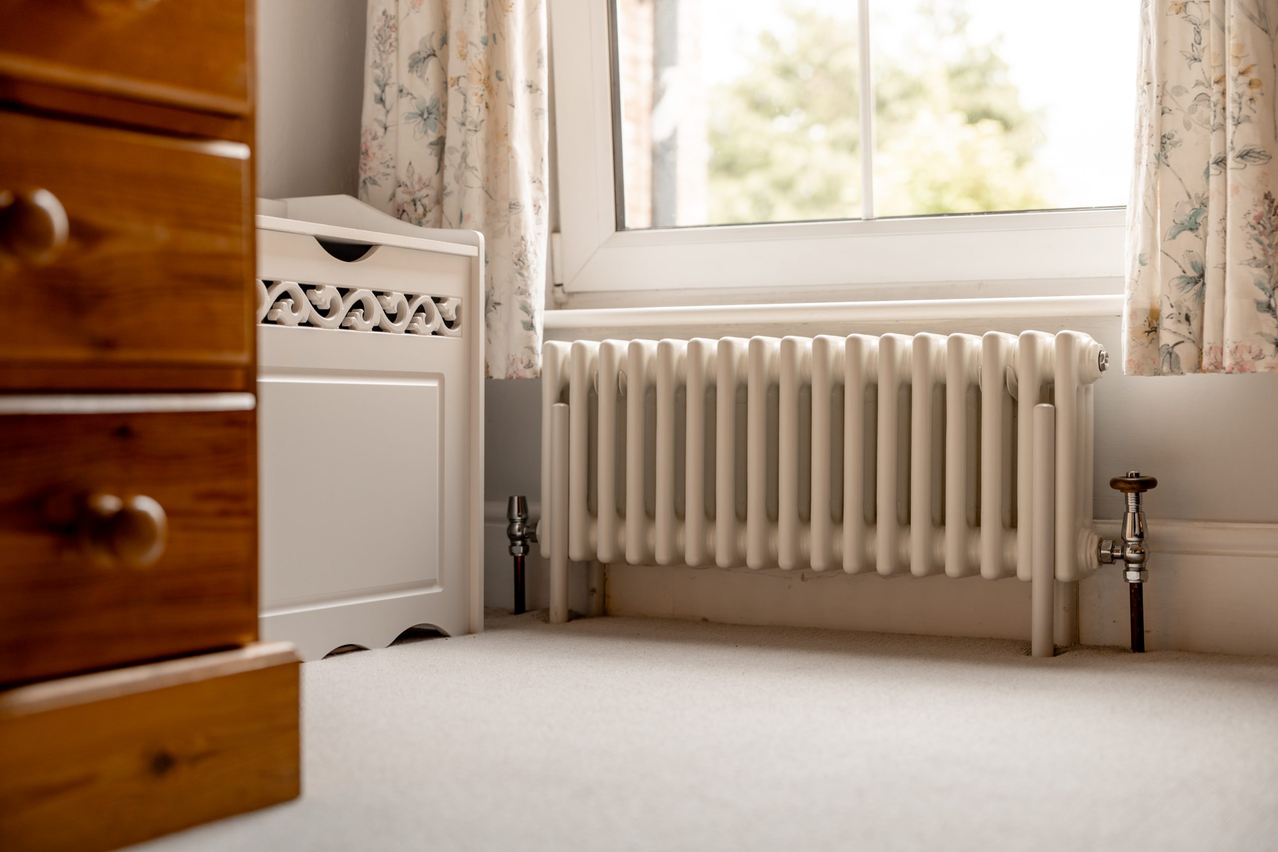 A white radiator beneath a window with floral curtains, next to a wooden chest of drawers and a white storage bench, on a light carpeted floor in a cozy room.