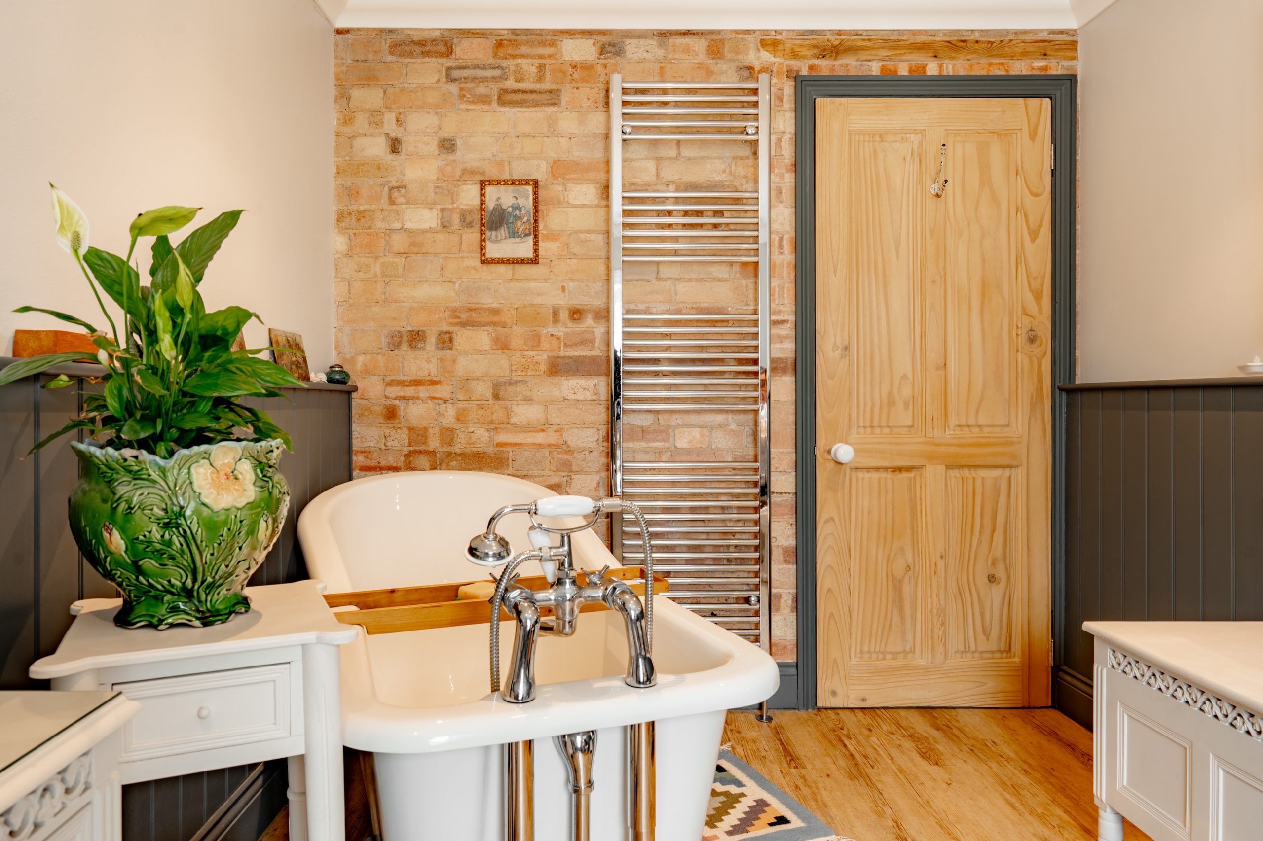 A cozy bathroom with a freestanding white bathtub, a wooden door, exposed brick wall, towel radiator, green leafy plant in a decorative vase, and light wood floors.