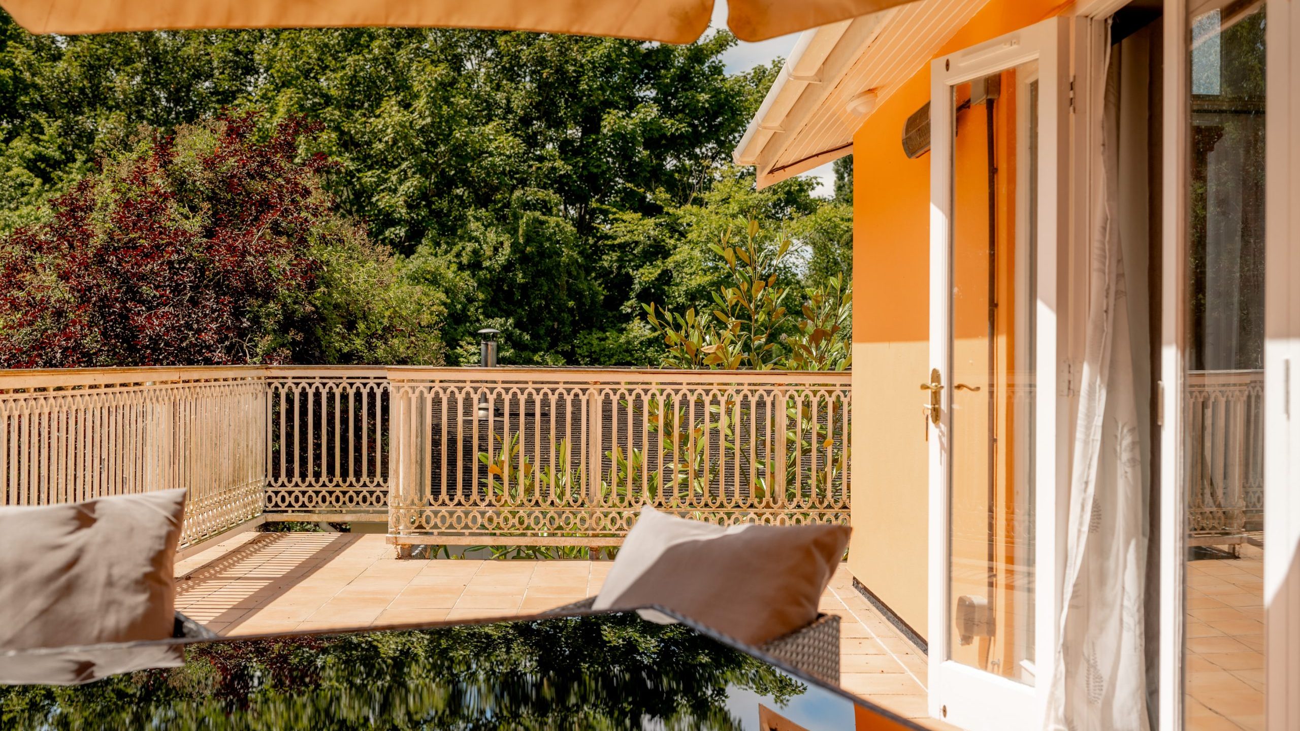 A sunny terrace with a table and cushions, light orange walls, an open glass door, a metal railing, and lush green trees in the background under a large beige umbrella.