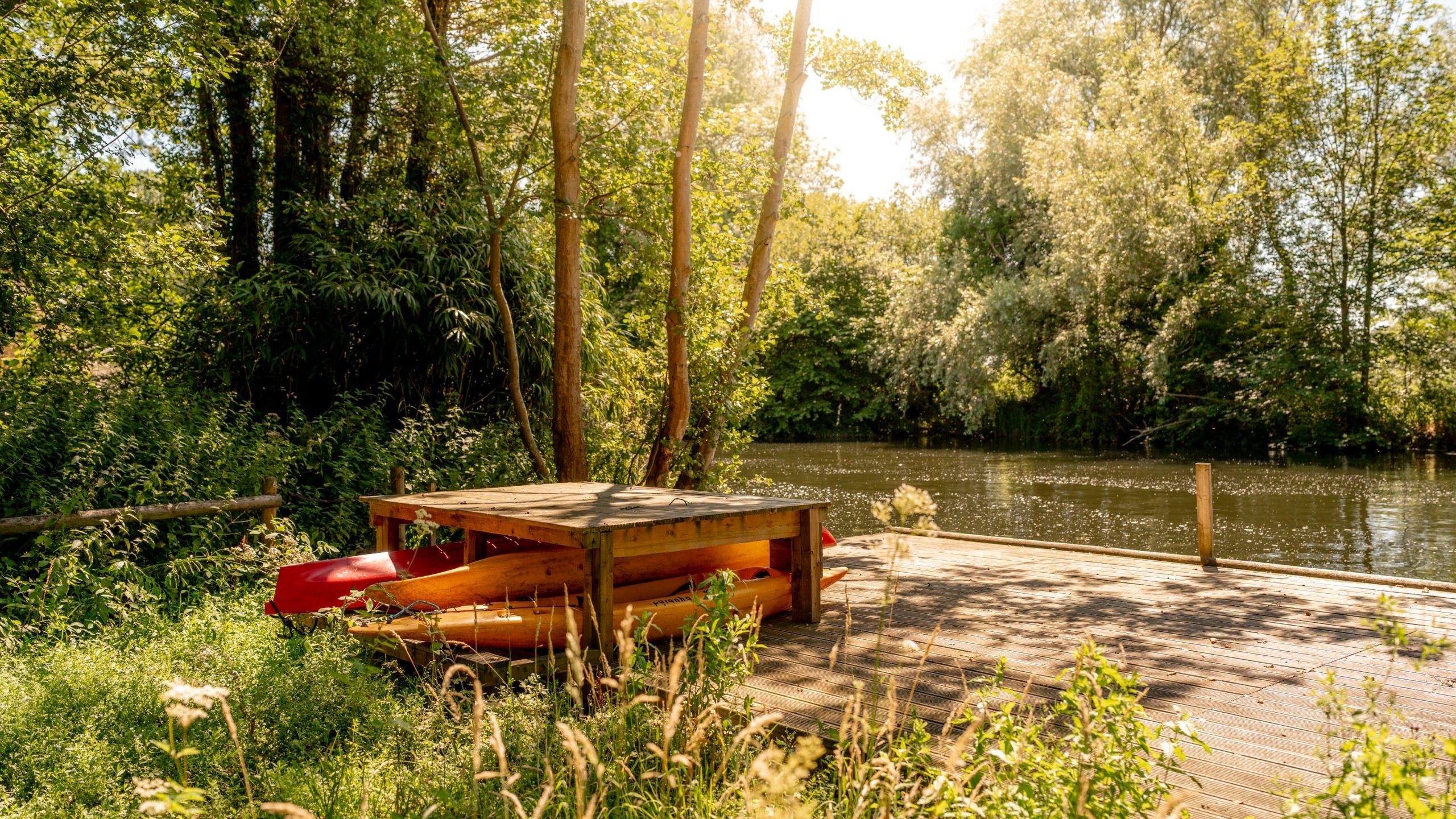 A wooden picnic table sits on a sunny dock by a calm river, surrounded by lush green trees and tall grass. A red kayak is stored underneath the table. Sunlight filters through the leaves, creating a peaceful scene.